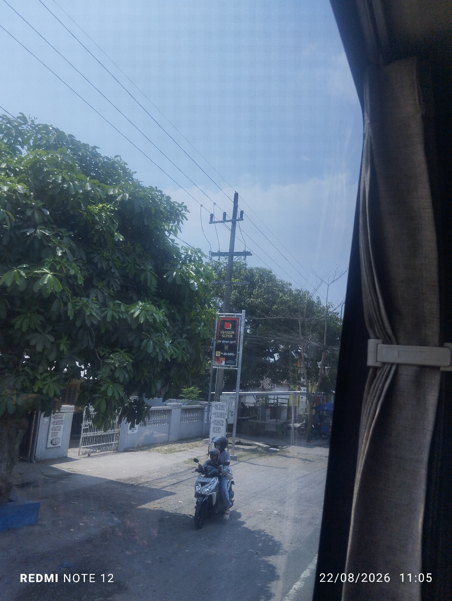 The image captures a street scene in Jiwan, Indonesia, on August 22, 2026, at 11:05 AM. The perspective is from inside a vehicle, looking outwards through a window partially obscured by a curtain. A large, leafy green tree dominates the left side of the frame, casting shadows on a paved road. In the mid-ground, a person wearing a helmet is riding a motorcycle. To the right of the motorcycle, a tall utility pole with electrical wires stretches across the sky. Further into the background, there are buildings and more trees. A sign is visible on the right side of the street, featuring text and an image. The sky above is clear and blue with some wispy clouds. The bottom left corner of the image displays "REDMI NOTE 12".
