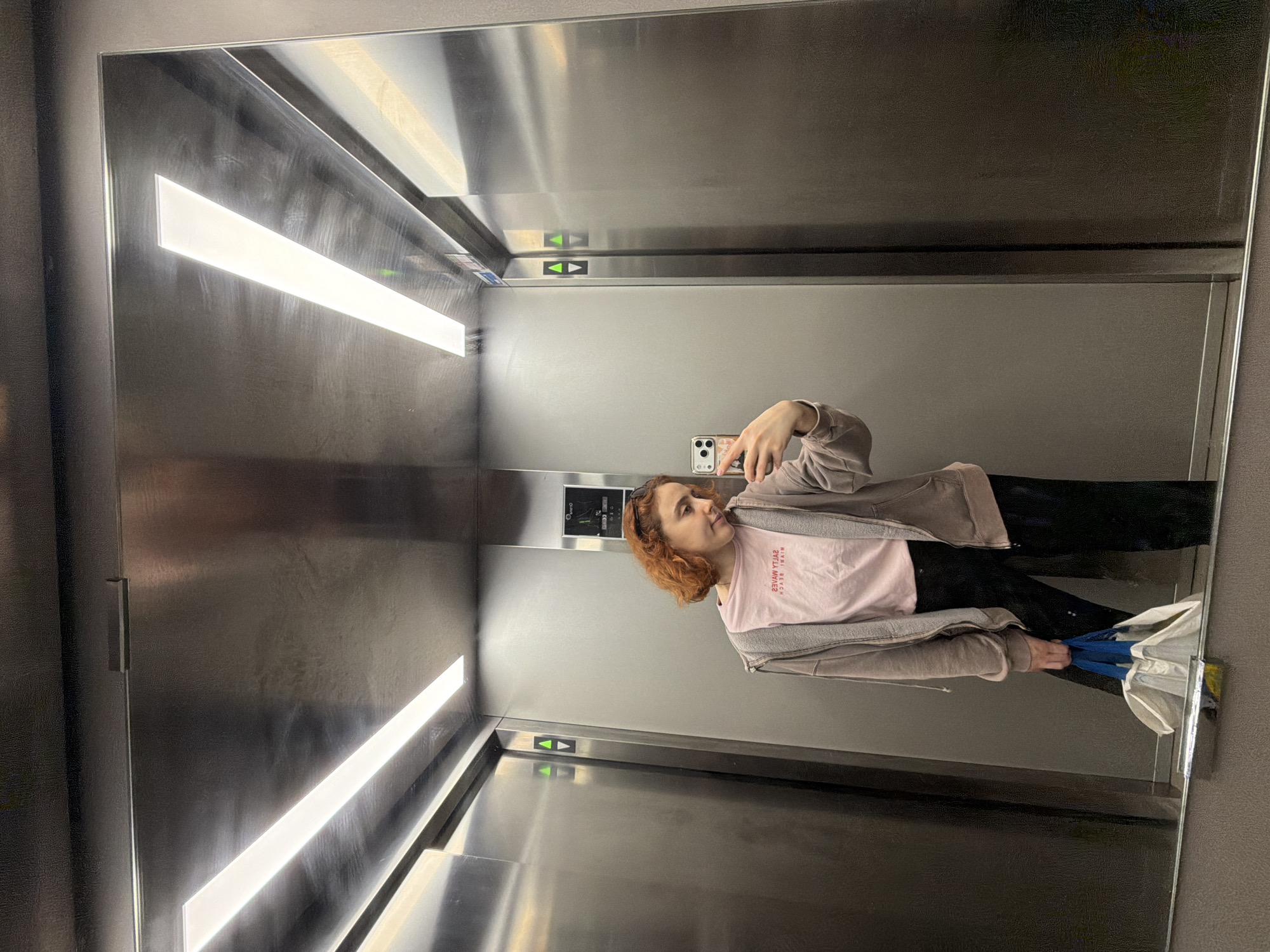 A young woman with short, reddish-brown, curly hair is taking a selfie in a mirrored elevator. She is wearing a light pink t-shirt with red lettering and a gray zip-up hoodie over it, along with black pants. In her right hand, she holds a smartphone, and in her left hand, she carries a white and blue tote bag. The elevator interior is constructed with brushed stainless steel panels, and the ceiling features two long, rectangular LED light fixtures. The woman is positioned in the center of the frame, with elevator doors visible on either side and directional arrows indicating upward movement. There are no discernible landmarks or clues to suggest the location is specifically in Strasbourg, France. The overall scene is a typical reflection within an elevator.