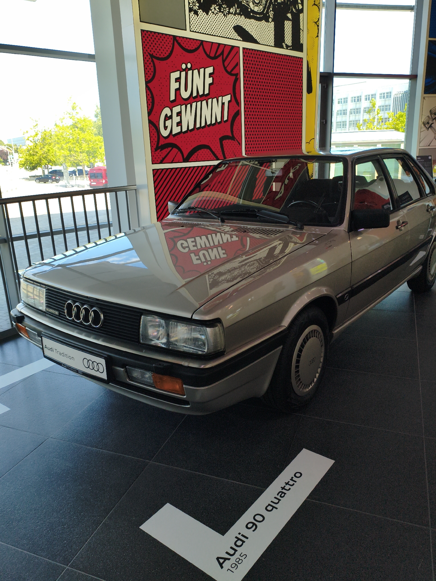 A silver Audi 90 Quattro, model year 1985, is displayed indoors on a dark tiled floor. The car is a four-door sedan with its front end facing the viewer's left and slightly towards the camera. It features the distinctive Audi rings emblem on the front grille and a license plate that reads "Audi Tradition." The interior of the car is visible through the windows, showing black seats and a steering wheel. Reflections on the car's polished surface reveal the surrounding environment, including what appears to be a large window with a view of trees and buildings outside, and a colorful graphic art installation featuring red and white pop art lettering that reads "FÜNF GEWINNT." In the background, a railing separates the display area from what looks like a public walkway or street. A white line on the floor points towards the car, with text that reads "Audi 90 quattro 1985." The setting is likely the Audi Forum in Ingolstadt, Germany, an exhibition space showcasing Audi's history and models. No people are present in the immediate vicinity of the car.