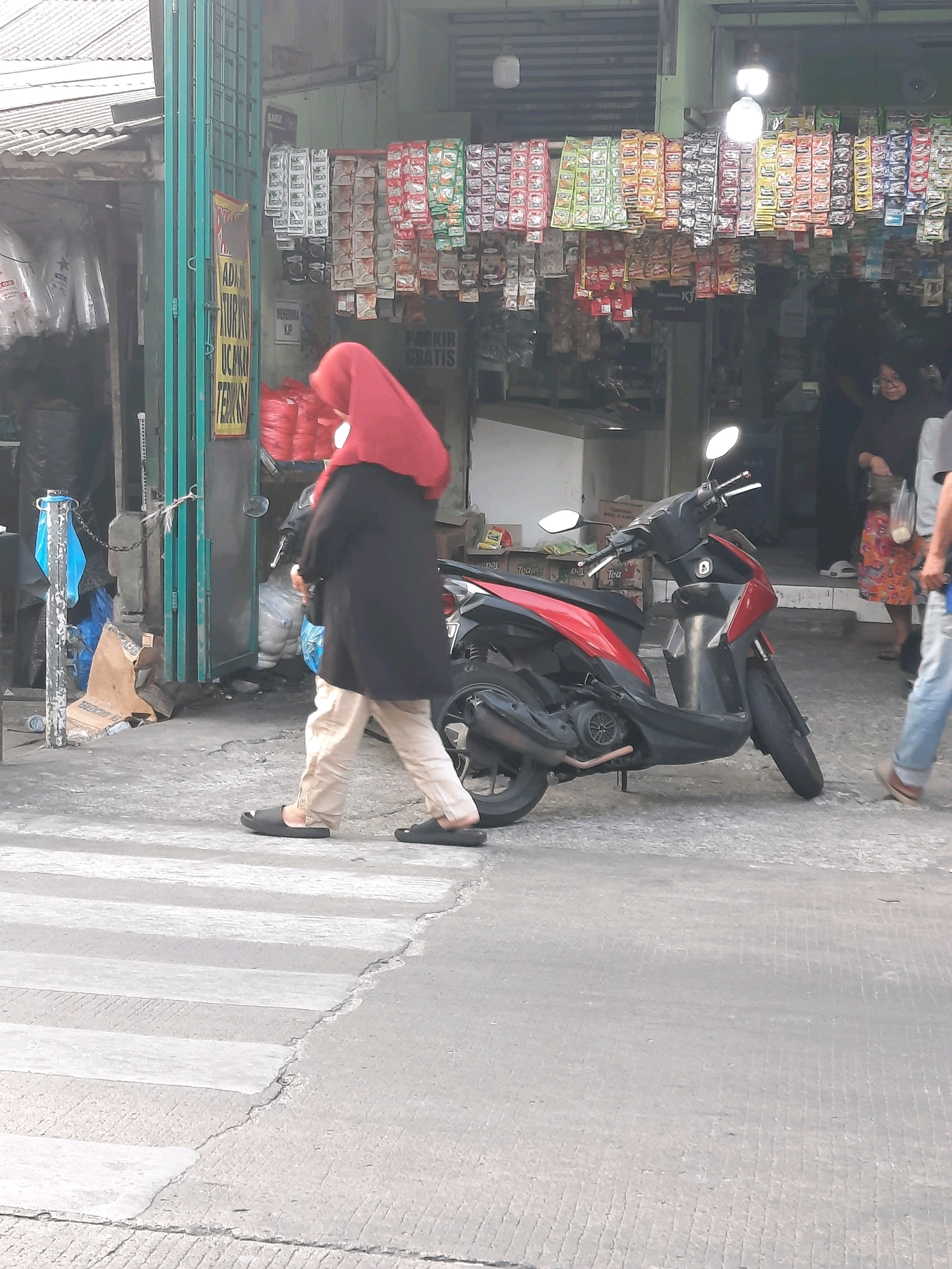 A woman wearing a red hijab and a black outer garment walks across a zebra crossing in front of a storefront in East Jakarta, Indonesia. She is wearing light-colored pants and dark sandals. To her right, a red and black scooter is parked on the sidewalk. The storefront displays numerous small packages of products on shelves and hanging racks. Behind the scooter, other individuals are visible, including a person in a patterned skirt and another person in jeans. The ground in front of the storefront is paved, and there are various items on the sidewalk, including bags and a cardboard box. The overall scene depicts a typical street view of a commercial area.