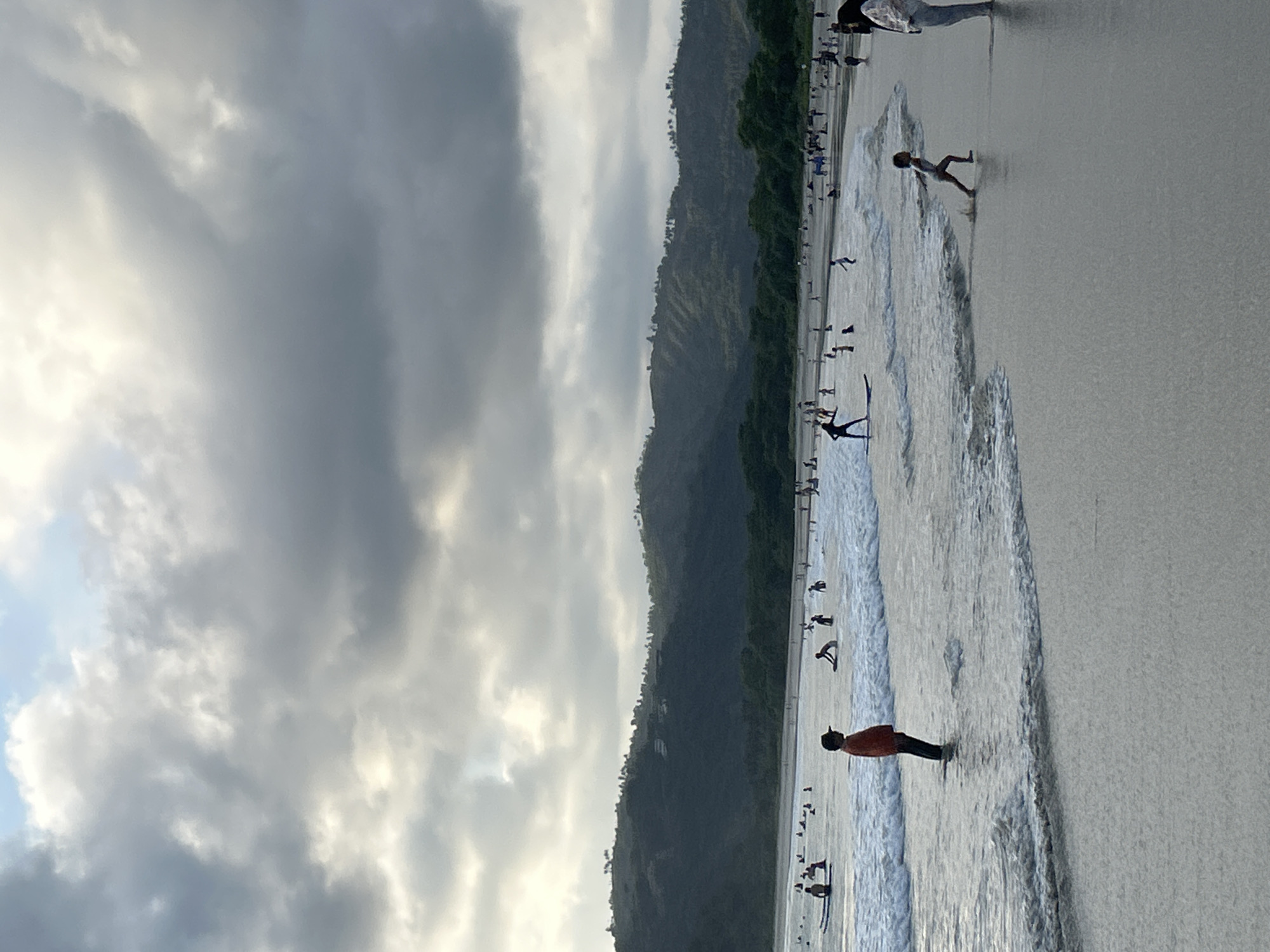 The image depicts a crowded beach scene at Selong Belanak, Indonesia, under a dramatic, cloudy sky. In the foreground, a young boy wearing an orange shirt and a cap stands in shallow water. To the right, a child runs along the wet sand. In the middle ground, numerous people are scattered across the water and beach. Several individuals are on surfboards, some actively surfing while others wait for waves. The water has moderate waves breaking near the shore. The background features a densely vegetated, hilly landscape that slopes down towards the sea. The sky is filled with heavy, gray clouds, with some brighter patches suggesting the sun attempting to break through. The overall atmosphere is that of a busy recreational day at the beach.