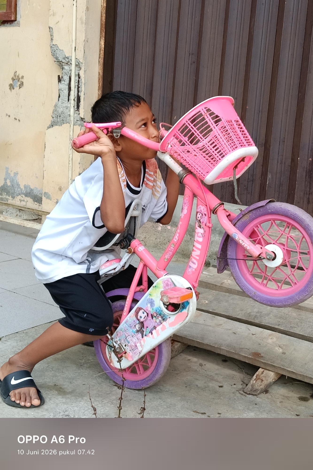 A young boy, appearing to be of Indonesian ethnicity, is kneeling beside a pink bicycle with a white basket. He is wearing a white t-shirt with black trim and black shorts, and black Nike slides. His left hand is on the handlebars, and he is looking upwards and to his right. The bicycle is decorated with cartoon characters and has pink wheels. The scene appears to be outdoors, with a textured wall and a wooden door in the background. The ground is paved with concrete, and there are some dried plant stems on the ground. The image also includes a timestamp at the bottom indicating "OPPO A6 Pro 10 Juni 2026 pukul 07.42," suggesting it was taken in Lhokseumawe, Indonesia on June 10, 2026, at 7:42 AM. The overall impression is that of a candid moment captured in a residential or urban outdoor setting.