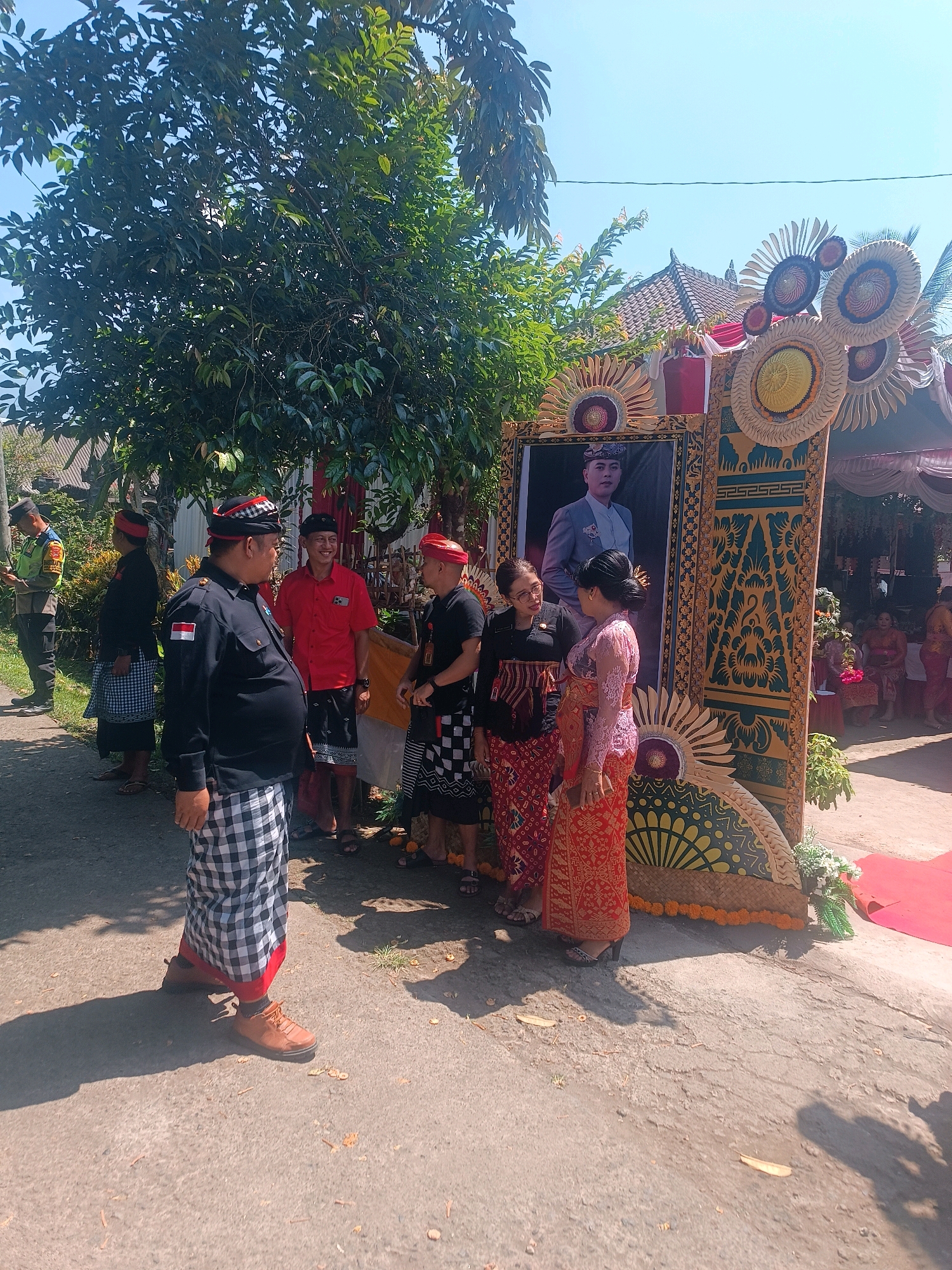 The image depicts an outdoor gathering in Penebel, Indonesia, likely a ceremony or celebration. In the foreground, a man in a black uniform with a red and white armband, a black cap, and a checkered sarong stands facing to the left, talking to another man. To his right, a man in a red shirt and a checkered sarong is looking towards the left and smiling. Two women are standing next to a large framed portrait, looking at each other. The woman on the left wears glasses and a dark top with a red patterned sarong, while the woman on the right wears a pink lace top and an orange patterned sarong. The portrait features a young man in a suit. The background reveals a decorated structure with circular, fan-like ornaments and intricate carvings, likely part of a stage or entrance. A red carpet is visible to the right. Several other individuals are present in the scene, some standing and others seated under a canopy in the background. Lush green trees and a clear blue sky dominate the upper portion of the image. The ground is paved with asphalt and scattered with fallen leaves.