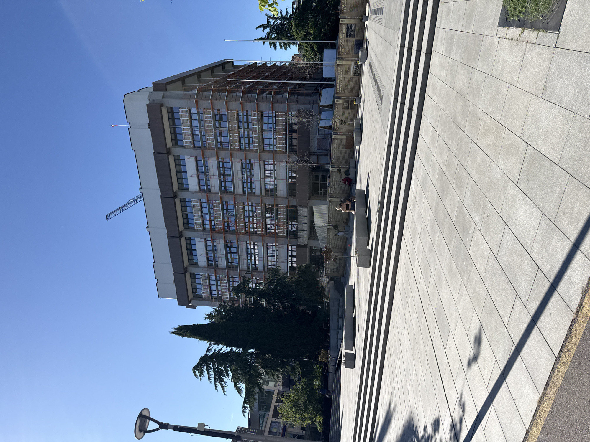 The image shows the Regional Administration building in Stara Zagora, Bulgaria. The building is a large, multi-story structure with many windows and scaffolding. A crane is also visible near the top of the building. In front of the building is a large paved area with steps leading up to it. There are people sitting and walking in this area. There are also trees and lampposts in the foreground. The sky is clear and blue. Close to the building, signs with text in Bulgarian can be seen.
