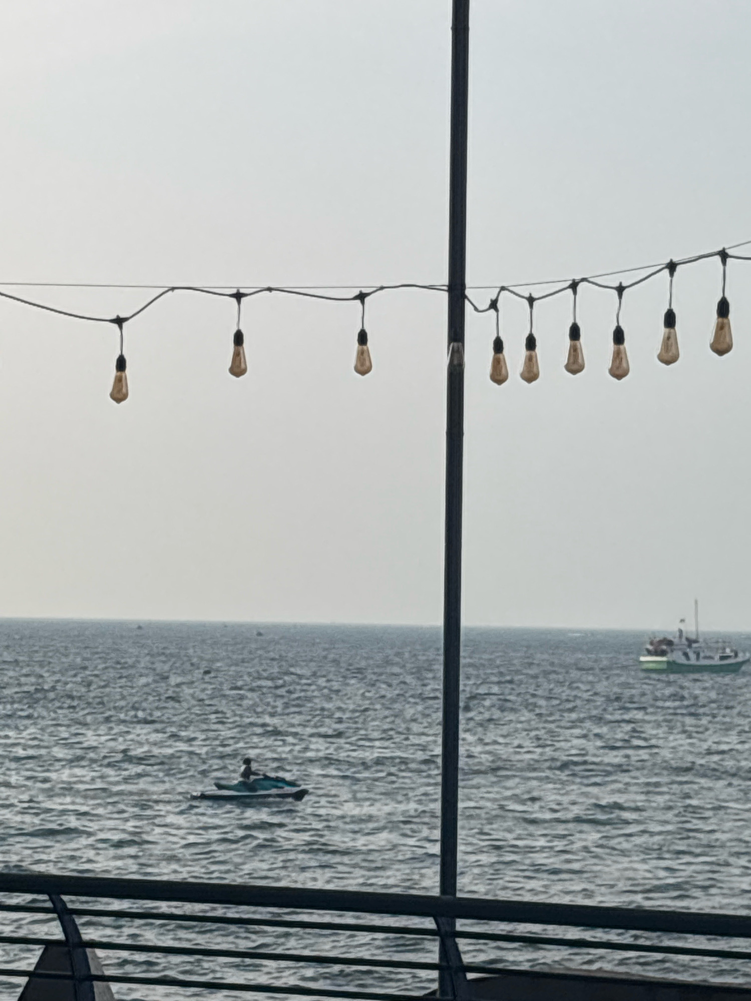 This is a photograph taken from a slightly elevated position overlooking the ocean in North Jakarta, Indonesia. In the foreground, a dark railing runs horizontally across the bottom of the frame, with a thin black pole rising vertically on the right side. Above the railing, a string of bare Edison-style light bulbs hangs from a wire, stretching across the upper portion of the image against a hazy sky.
The ocean fills the midground, its surface rippled with waves. On the left side of the ocean, a person is visible riding a jet ski. Further in the distance on the right side of the ocean, a white and green boat is moving across the water. The horizon line is visible where the ocean meets the sky, and the sky itself is overcast and light in color. The overall impression is of a waterfront scene on a cloudy day.