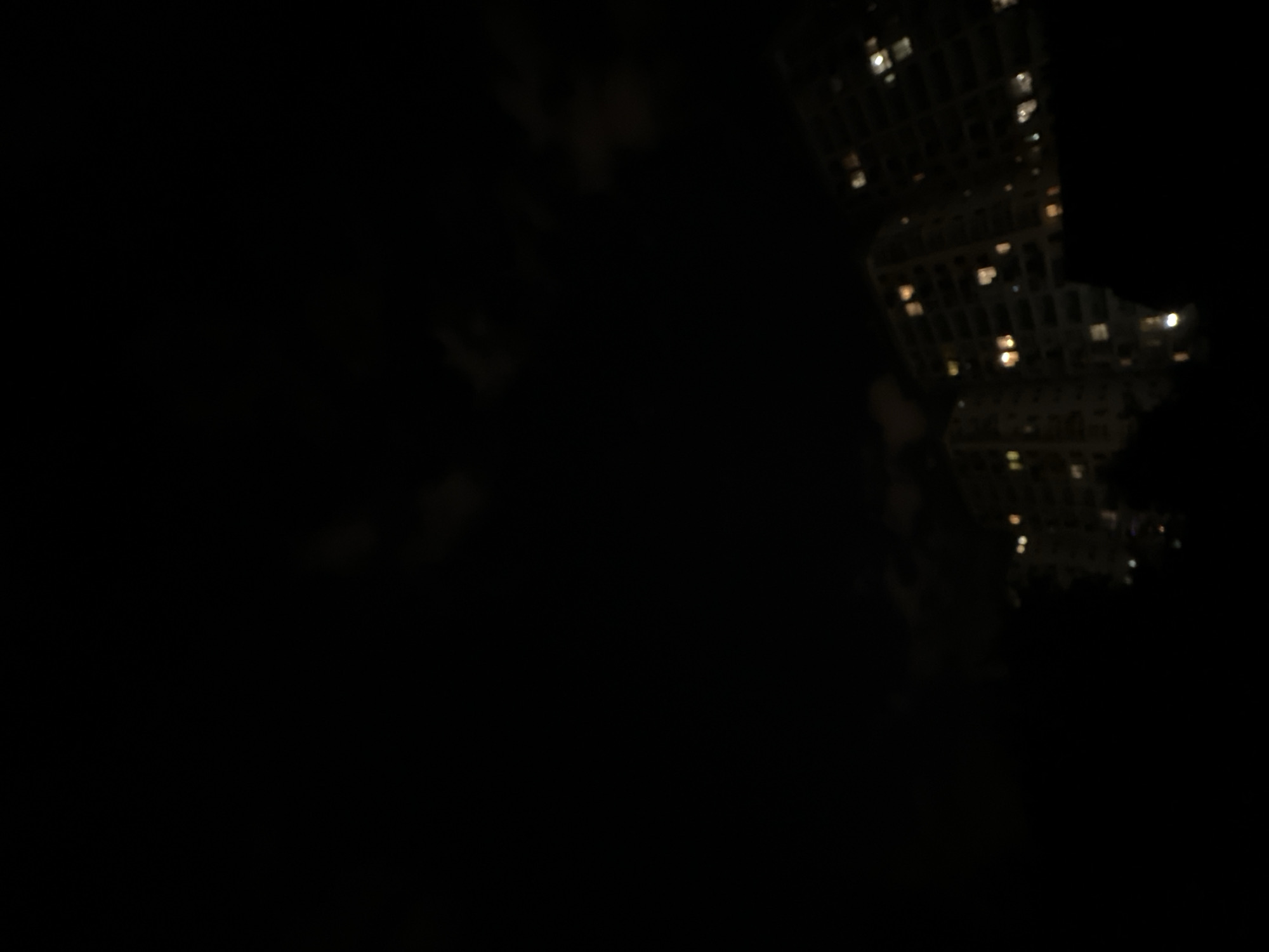 The image shows a night scene in Minsk, Belarus. The view is mostly dark, but in the upper-right portion of the frame, there is a faint and distorted reflection of a building with illuminated windows, indicating an urban setting. The rest of the image is obscured by darkness, with faint outlines suggesting the presence of foliage or other dark objects.