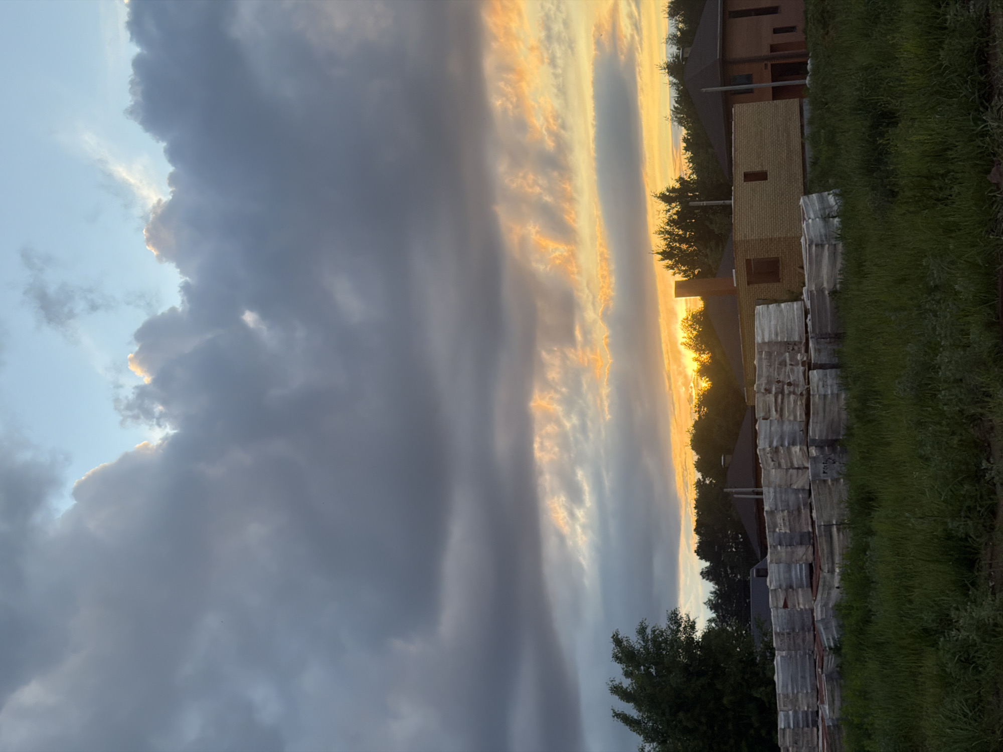 The image captures a sky with contrasting weather conditions. On the left side, there are heavy, dark gray storm clouds, while the right side shows a sunset with golden light filtering through lighter clouds. The sun's rays illuminate the clouds and create a striking contrast.
In the lower right corner, there's a glimpse of land with green grass and a partially constructed brick building. Stacks of wrapped construction materials are visible near the building. Trees stand in the background, silhouetted against the setting sun.
Given the reference to Chaikovsky, Russia, the location is likely within or on the outskirts of the city, possibly in a developing area where construction is underway. There are no people in the image. The dominant subjects are the contrasting weather conditions and the construction site at sunset.