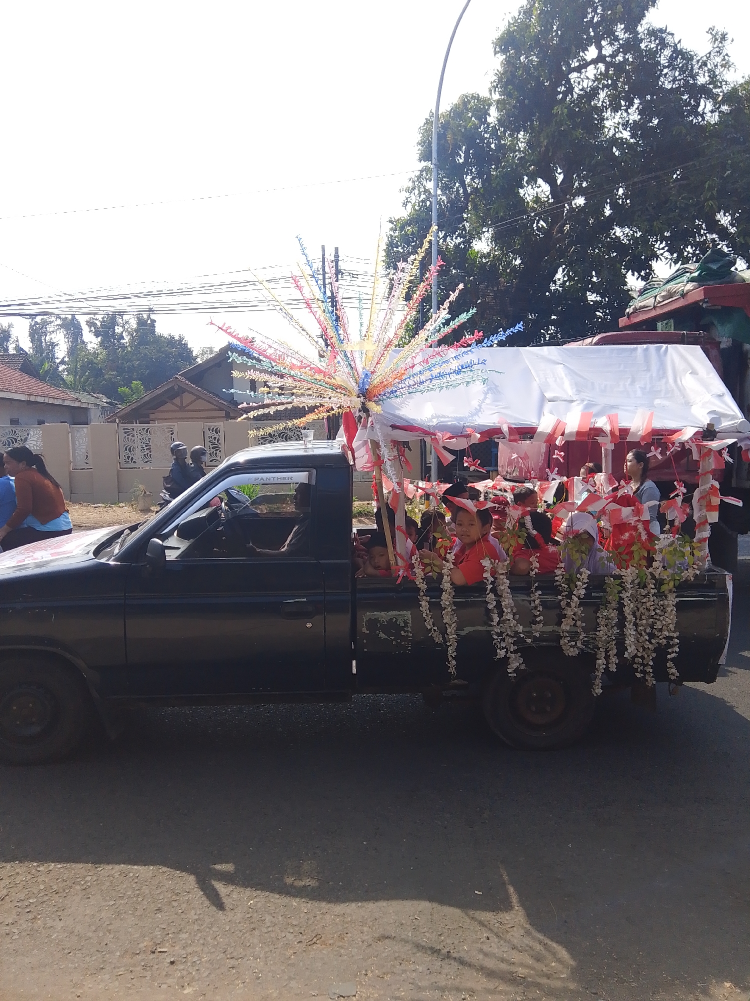 In the city of Kalinyamatan, Indonesia, a black pickup truck with a white canopy is decorated for a parade. The canopy is adorned with red and white Indonesian flags. Numerous children are seated in the truck bed, many wearing red shirts, and some are holding small Indonesian flags. The truck is decorated with colorful streamers emanating from the front and white floral garlands hanging from the sides. A woman in an orange shirt and a woman in a blue shirt are standing near the front of the truck. A man is visible in the driver's seat. In the background, houses and trees are visible under a bright sky. The truck is on an asphalt road, with shadows of trees and objects cast on the road.