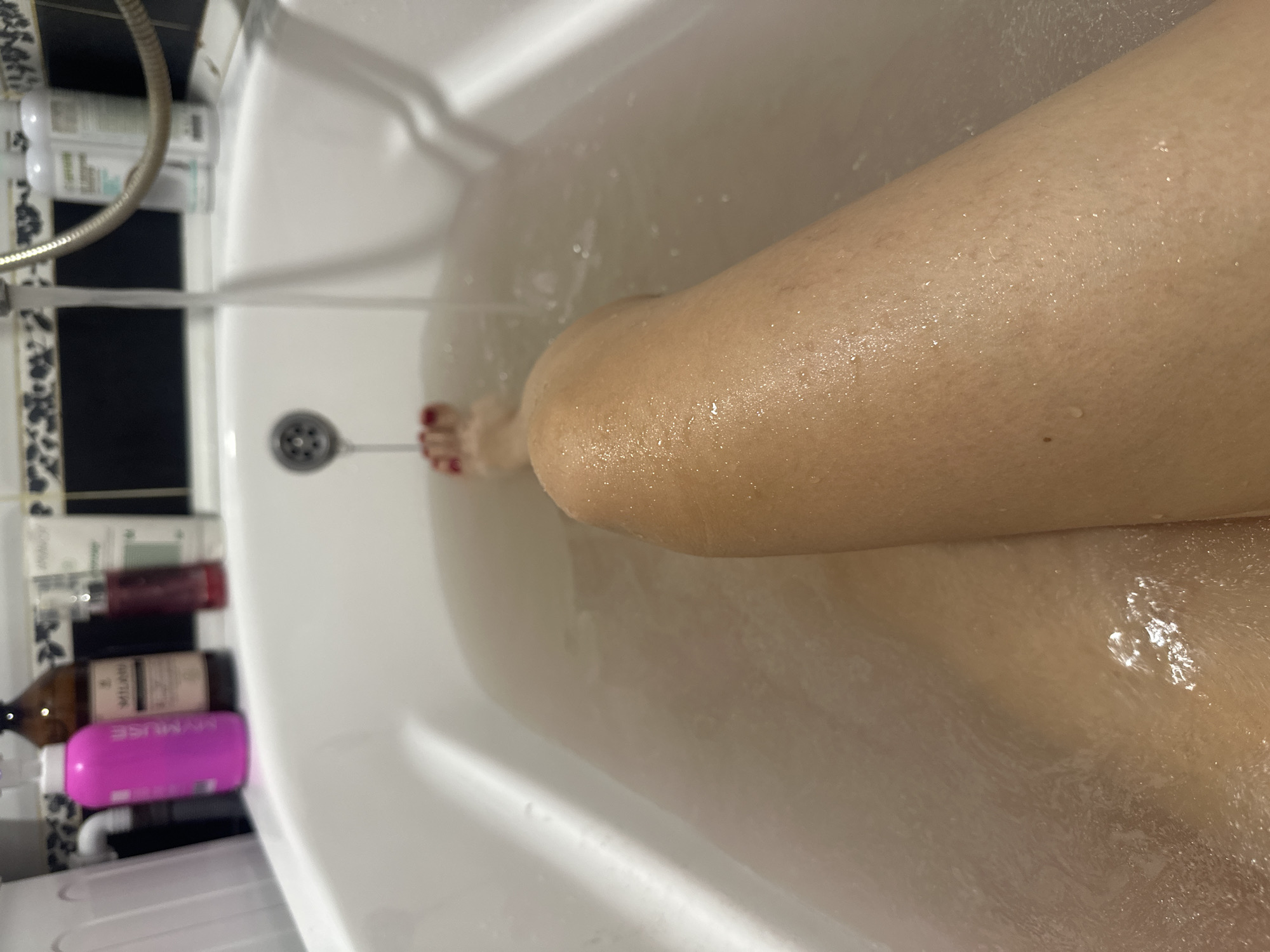 The image shows a close-up view of a person's legs and feet submerged in bathwater within a white bathtub. The water appears slightly murky. A drain is visible in the tub. On the edge of the bathtub, several bottles of toiletries are present, including a bright pink bottle, a dark amber bottle, and two white bottles. A chrome faucet is also visible, with water running from it. The background shows a tiled wall. The scene is indoors and appears to be a bathroom. The location in the city of Novoaltaysk, Russia cannot be determined from the image alone.