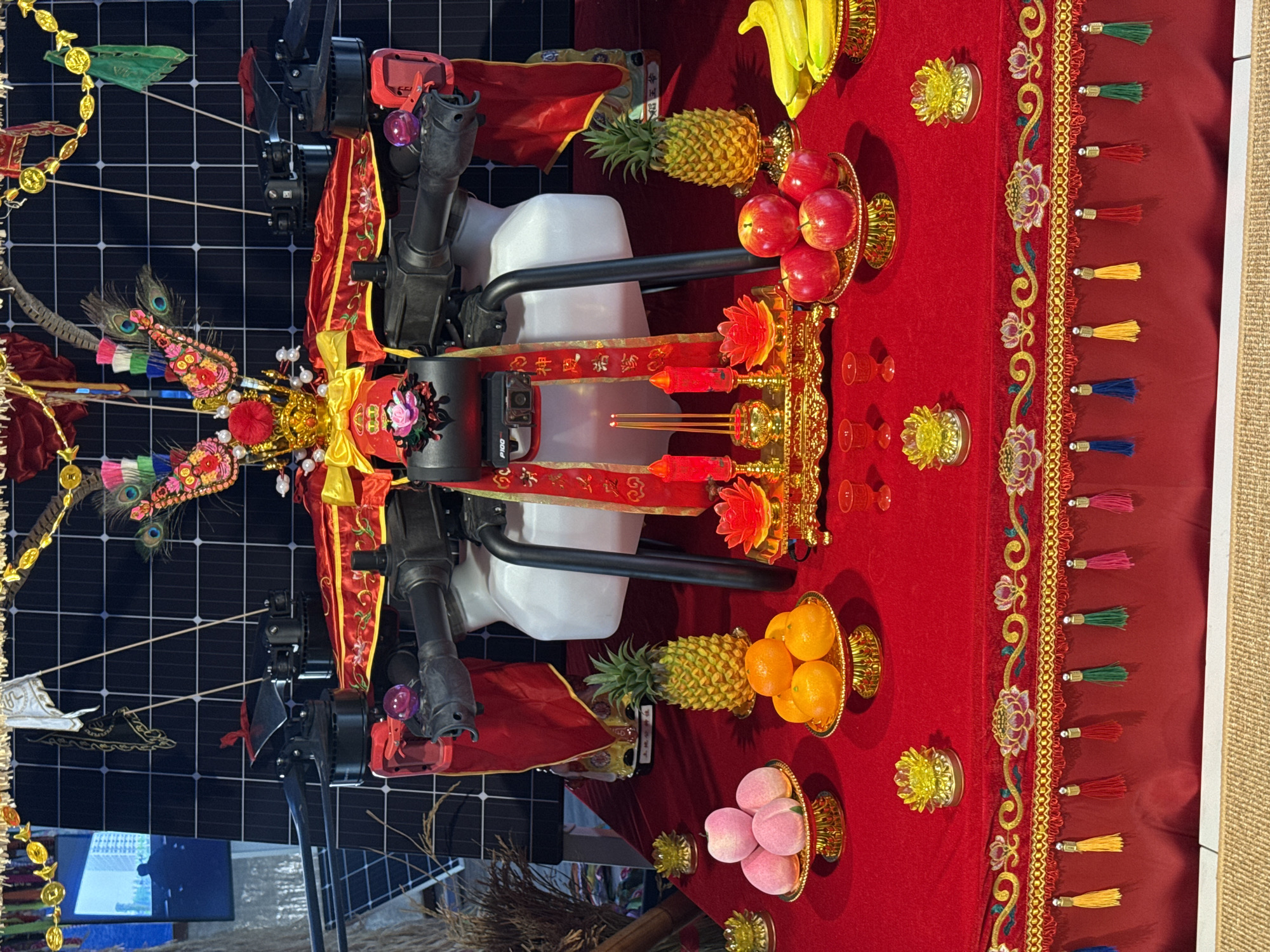 The image depicts a shrine-like arrangement set up on a red tablecloth with gold-colored tassels and embroidered designs along the edge. The arrangement includes various fruits displayed on ornate, golden stands: bananas, pineapples, apples, oranges, and peaches. There are also decorative golden objects, candles, and incense sticks.
At the center of the arrangement is a black and white machine-like object adorned with colorful decorations, peacock feathers, red fabric with gold trim, and banners with Chinese characters. The object appears to be the focus of the display.
The backdrop consists of a grid-like pattern, possibly a solar panel, and a small section of what appears to be a television screen. A broom is present near the bottom of the frame.
The location in Milan, Italy is not apparent from the visual information alone. There are no discernible elements within the image to confirm its geographical location within Milan.
