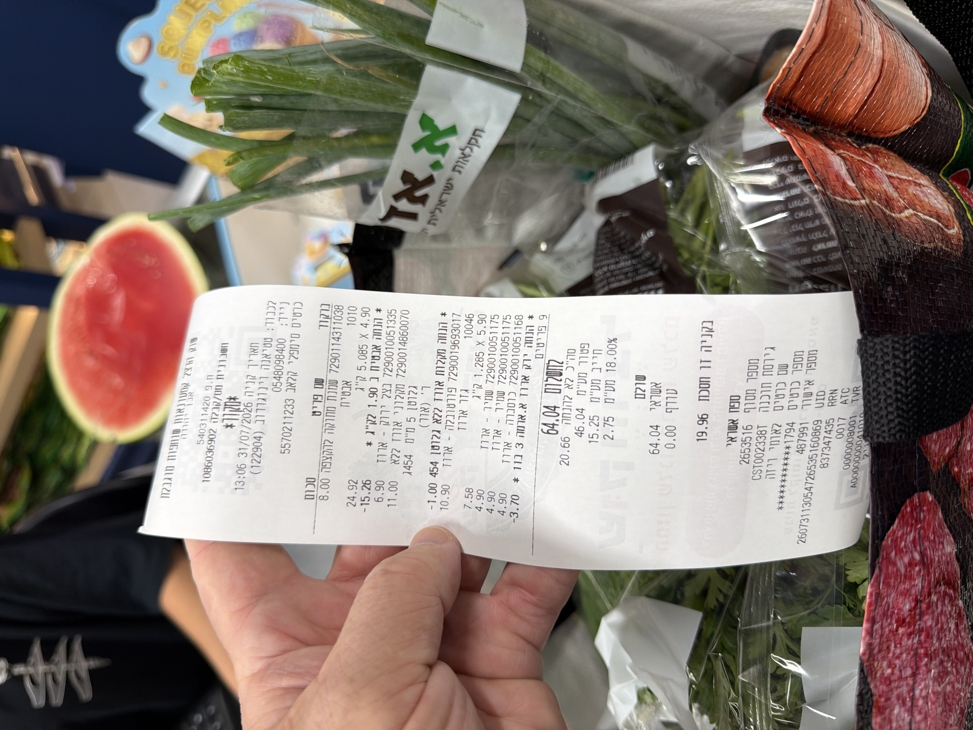 The image shows a close-up of a grocery receipt held by a person's hand, with a blurred background that includes a watermelon slice and green onions in plastic packaging. The receipt is in Hebrew and details various items purchased, including their prices and quantities. The receipt also indicates the location of purchase as "Even Yehuda, Israel," with a date of "31/07/2026." The overall scene appears to be inside a supermarket or market.