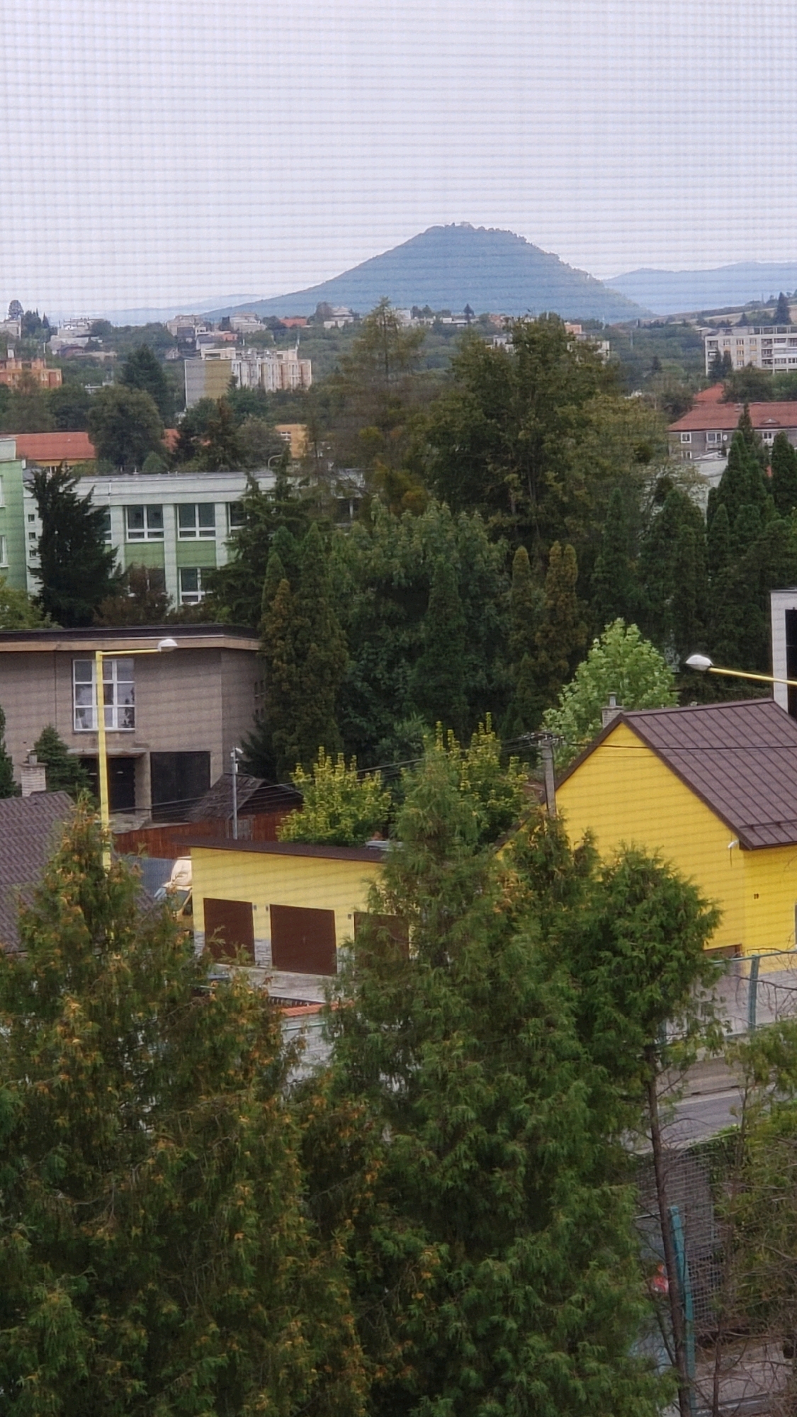 The image depicts a cityscape with a dominant hill or mountain in the background. In the foreground and midground, various buildings and trees are visible. There are residential buildings, some with multiple stories, and smaller structures that appear to be garages or sheds. The vegetation consists of numerous trees, including coniferous and deciduous varieties, in various states of foliage. The overall impression is that of a suburban or semi-urban environment on a cloudy day.
Based on a visual analysis of the image's characteristic landscape, specifically the distinctive conical shape of the dominant hill in the background, it is identifiable as the Zámčisko hill. Zámčisko is a prominent geological formation located in the immediate vicinity of the city of Prešov, Slovakia. It is a well-known landmark and a common feature in many views of the city from various vantage points. The presence of this specific hill strongly suggests that the image was captured within or overlooking the city of Prešov.
