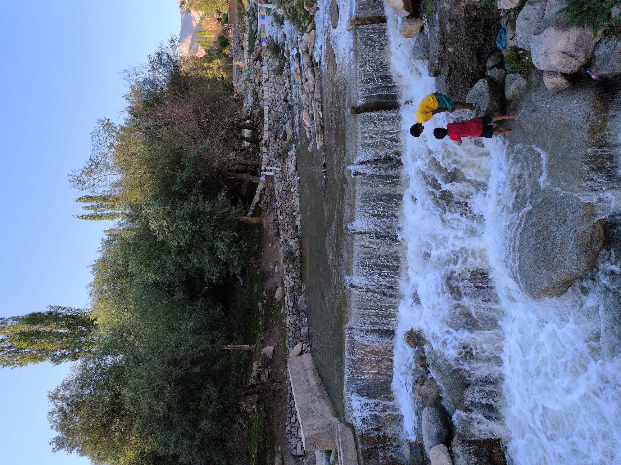 Two young boys are standing on rocks in a shallow river with a waterfall. One boy, wearing a yellow t-shirt and green shorts, is on a large rock at the edge of the waterfall. The other boy, wearing a red t-shirt and black shorts, is closer to the waterfall's edge, reaching towards the water. The river flows over a man-made concrete structure that creates the waterfall. The background shows lush green trees, rocky terrain, and a clear blue sky. In the distance, mountains are visible. Prayer flags are strung across the upper part of the scene, indicating a spiritual or cultural context. The overall environment suggests a natural landscape with some man-made elements, likely in a mountainous region.