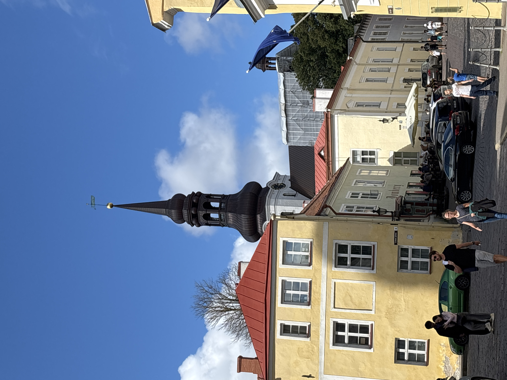 The image depicts a street scene in Tallinn, Estonia, likely in the Old Town. In the foreground, a cobblestone street is visible with several people walking. To the left, a yellow building with a red roof is prominent, featuring several windows. Behind this, a tall, dark-colored church spire dominates the skyline, topped with a weather vane. Further back, other buildings with light-colored facades and red roofs are present, along with trees. A blue flag with stars is visible flying from a building on the right side of the frame. The sky is bright blue with scattered white clouds. Several cars are parked or driving on the street. The overall impression is of a sunny day in a historic European city.