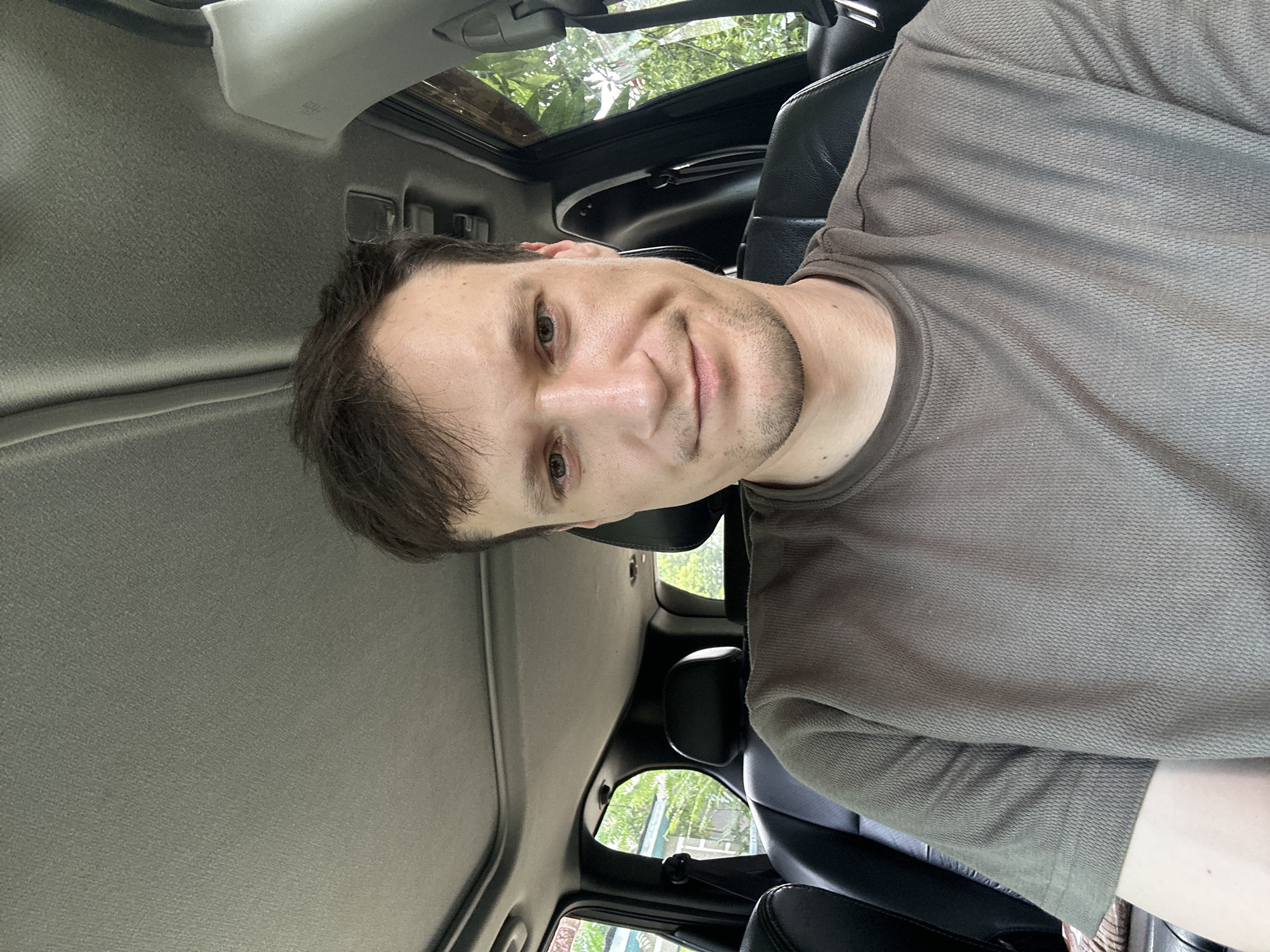 A man with short, dark hair and light skin is sitting in the driver's seat of a car. He is wearing a short-sleeved, olive-green t-shirt. The car has black leather seats and a sunroof that is open. The windows are down, and green foliage is visible outside. The man is looking at the camera with a slight smile. The location is in the city of Makiv, Ukraine.