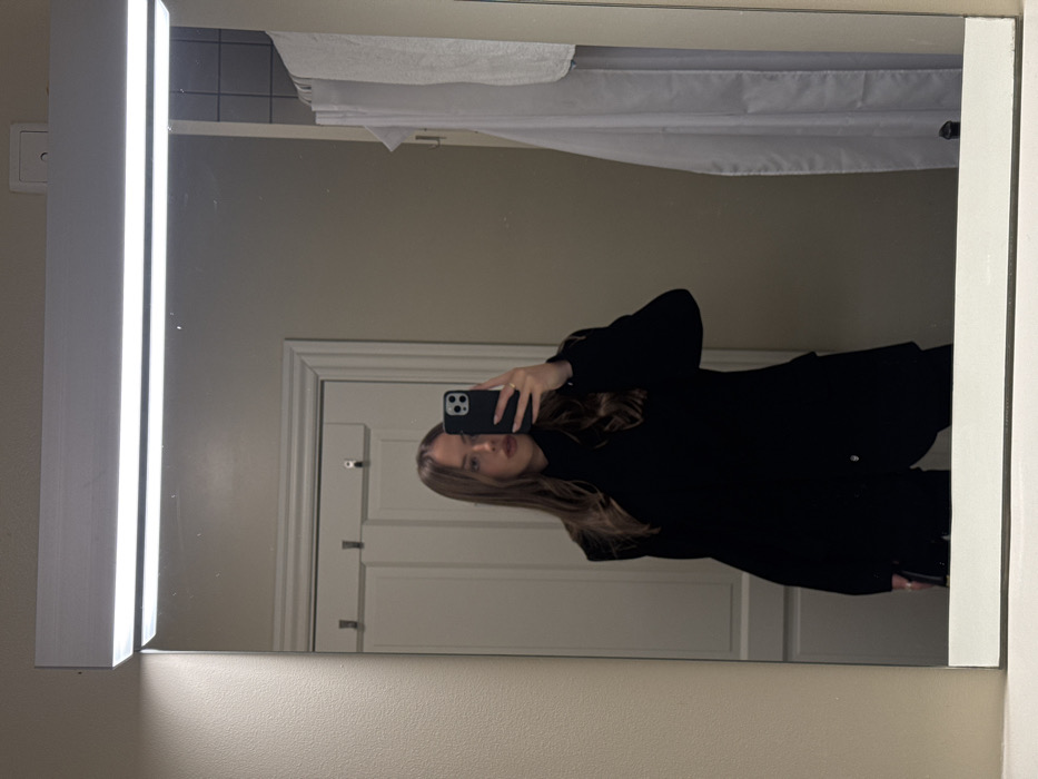 A mirror selfie shows a woman in a black coat holding a phone. She is looking directly at the camera. The mirror reflects a white door with silver handles and a hanging white towel above. A vertical light is on the left side of the mirror. The background is a beige wall.