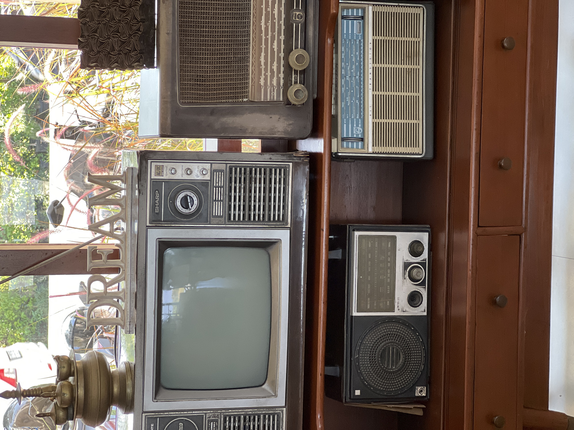 In the city of Ubud District, Indonesia, this image captures a shelf display of vintage electronic devices, including two radios and a television set, situated on a dark wooden furniture piece. Above the television, a metallic sculpture spells out the word "DREAM," positioned behind an ornate brass vessel. The background reveals a sunlit outdoor scene with greenery and what appears to be a motorcycle, suggesting a semi-outdoor or well-lit indoor setting. The items are arranged meticulously, creating a nostalgic and decorative arrangement.