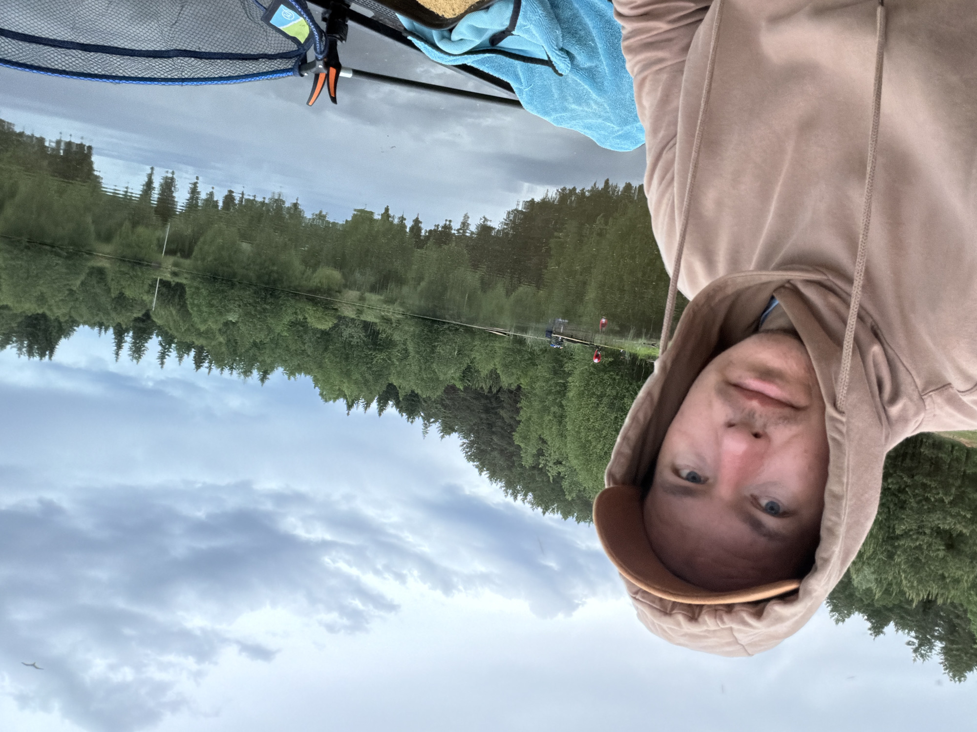 A selfie taken by a person wearing a beige hoodie and a brown cap. The background shows a body of water reflecting a treeline and cloudy sky, suggesting a location near a lake or river. On the left, portions of a landing net, fishing rod, and a blue towel are visible. This location is most likely in or near the city of Prikamskii, Russia.