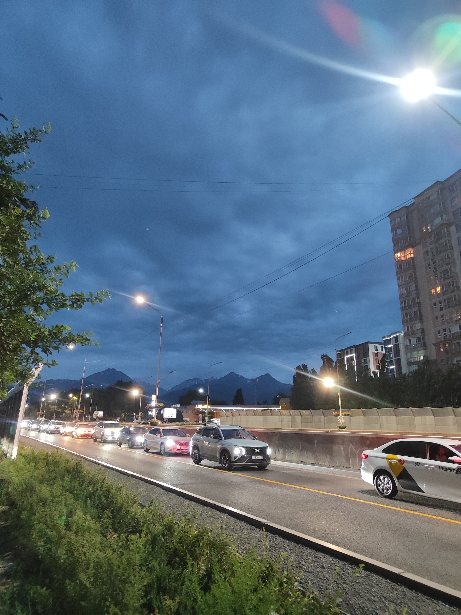 The image shows a view of a traffic jam on a road in Almaty, Kazakhstan, at dusk. Mountains are visible in the background, and several cars are lined up on the road, with their headlights on. A few streetlights are also visible, illuminating the scene. A tall apartment building is on the right side of the image. The sky is dark blue with some clouds. A white car with Yandex Taxi branding is in the foreground on the right.