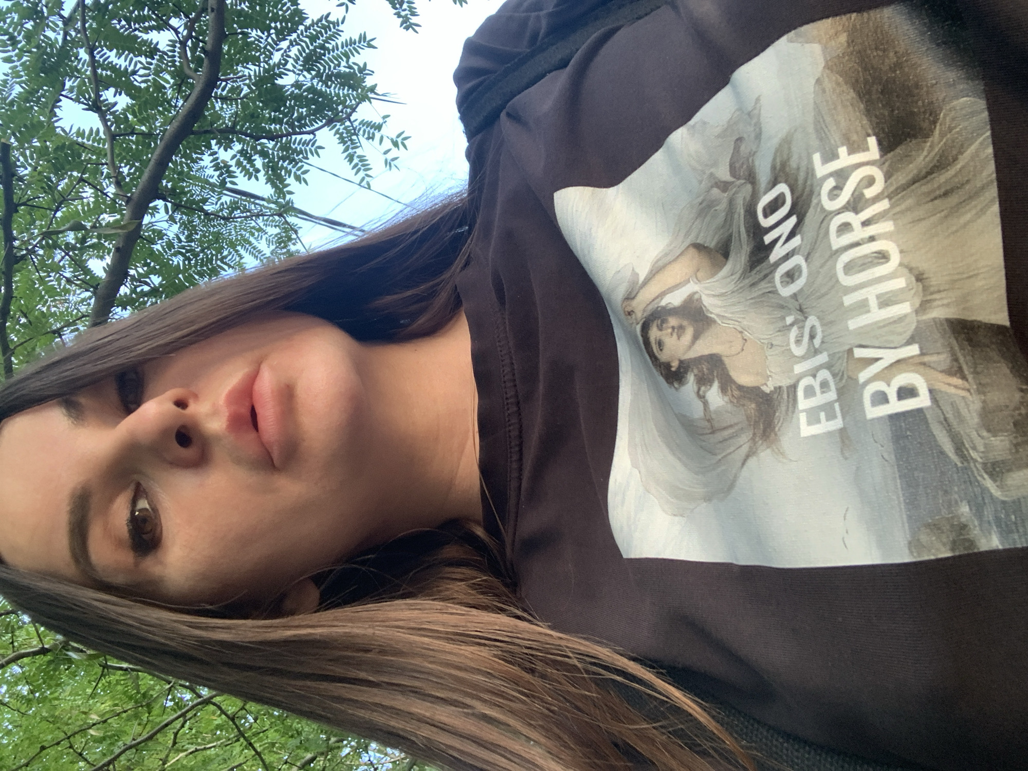 A low-angle, medium shot shows a woman from the chest up, looking slightly upwards and to the right of the camera. She has long, dark brown hair, brown eyes, and full lips. She is wearing a black t-shirt with a large, detailed image of a classical painting of a woman in a flowing white dress on a rocky outcrop overlooking the ocean. White text on the t-shirt reads "EBIS: ONO" and "BY HORSE." Green leaves and branches from a tree are visible in the upper part of the frame against a light blue sky. The woman is outdoors in an urban setting.