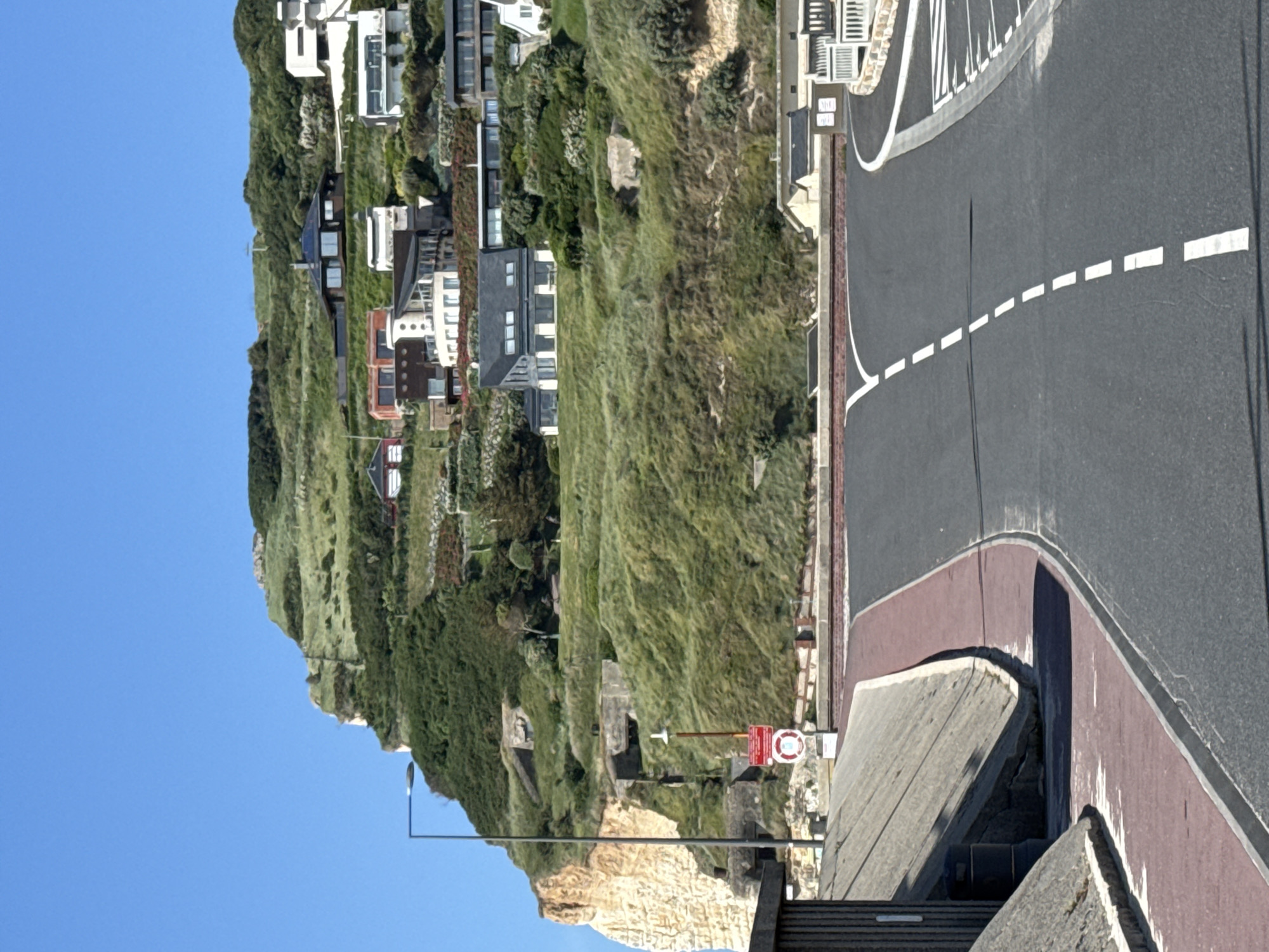 The image displays a bright, sunny coastal landscape, likely near Fécamp, France, featuring a road winding along a verdant hillside overlooking the sea.
In the foreground, a dark asphalt road curves gently, marked with white dashed lines on the left and a solid white line on the right. A reddish-brown paved shoulder or pathway runs alongside it. To the left of the road, a steep, lush green hillside rises, densely covered in grass and various shrubs and trees. Several multi-story houses of diverse architectural styles, from modern designs with large windows and balconies to more traditional buildings, are nestled into this verdant slope.
Towards the lower-left, a prominent light-colored, possibly chalk or limestone, cliff face is visible. Embedded within the lower section of the hillside or cliff are several concrete structures, resembling old bunkers or fortifications, one of which has a distinct rectangular opening. A black streetlight pole stands near the road at the bottom left. Near the road, a red circular sign features a white lifebuoy symbol with a red diagonal line through it, indicating a prohibition, though accompanying text is illegible. A smaller, rectangular sign is also visible on a pole nearby.
The sky is a clear, vibrant blue, indicating bright, sunny weather. The scene is brightly illuminated, suggesting it is midday or early afternoon. No people or active events are present, capturing a static and serene view of the coastal environment.