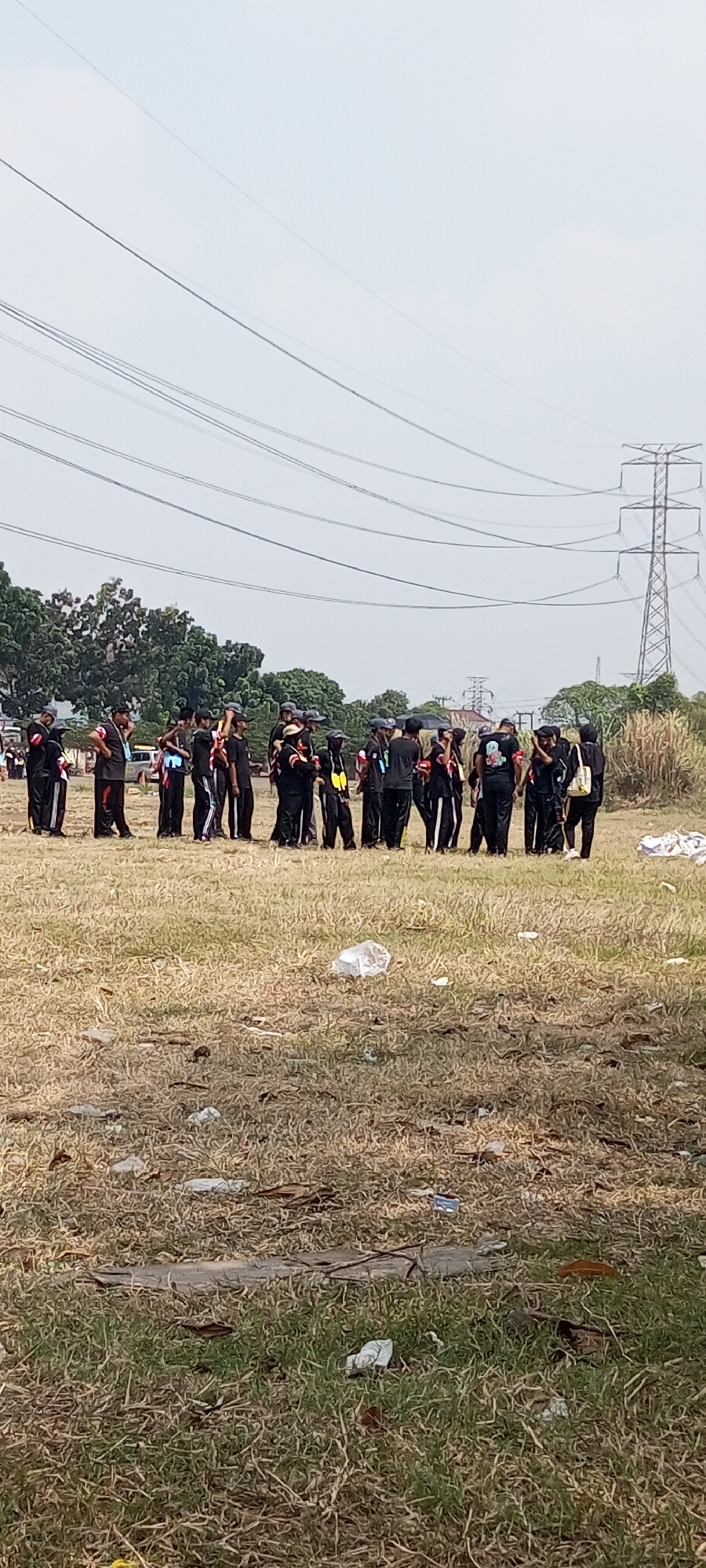 A group of approximately 20 young people, dressed in black uniforms with some red and white accents, are gathered in a dry, grassy field. They appear to be a team or group, possibly students, standing in a loose formation. Some individuals are holding items, and one person is holding what looks like a yellow bag. In the background, there are tall trees and a large electricity pylon with power lines stretching across the sky. The ground is littered with dried grass, leaves, and some pieces of trash, including white plastic bags. The overall environment suggests an outdoor gathering in a somewhat undeveloped or rural area. This scene is located in Lambangjaya, Indonesia, as indicated by the surrounding power lines and the general landscape typical of the region.