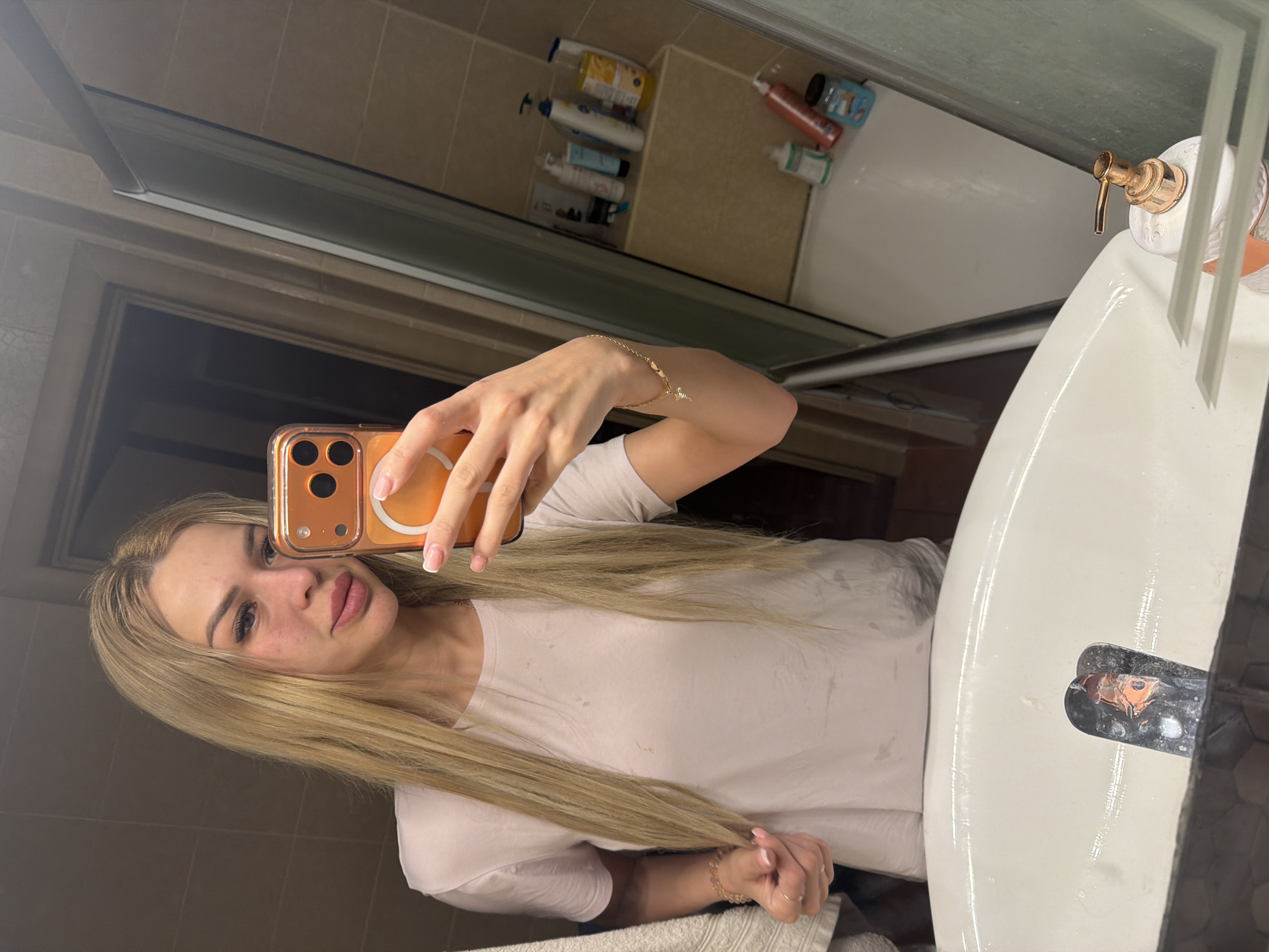 In a bathroom, likely in Kyiv, Ukraine, a woman with blonde hair is taking a mirror selfie. She is wearing a light pink t-shirt and has a bracelet on her left wrist. She is holding an orange phone to take the photo. Various toiletries are visible on a shelf in the background, including bottles and containers. The sink in the foreground shows some water and reflections.