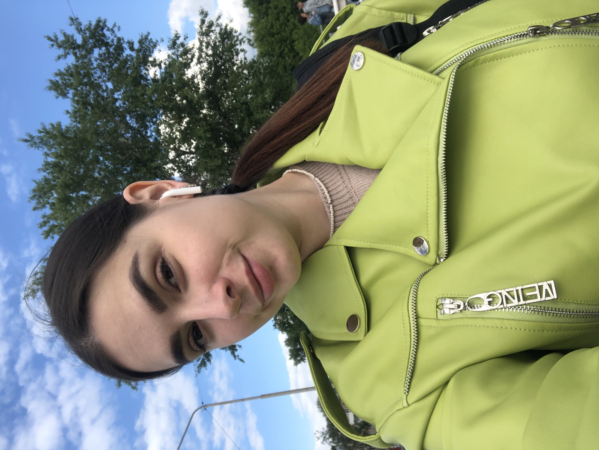 The image is a selfie, rotated 90 degrees counter-clockwise, featuring a young woman outdoors in Chaikovsky, Russia. The setting is a bright, sunny day with a clear blue sky and scattered white clouds. The background also includes the lush green foliage of deciduous trees and a visible section of a utility pole or street light.
The primary subject is a young woman with dark brown hair, styled with a side part and pulled back, revealing a section of a braid resting on her right shoulder. She has prominent dark eyebrows, dark eyes, and a subtle, soft smile on her lips, conveying a calm and pleasant demeanor. She is wearing a light-colored, possibly beige or light brown, ribbed knit top beneath a vibrant lime green faux leather jacket. A white wireless earbud is noticeable in her right ear, and a delicate, light-colored beaded necklace is visible around her neck.
A black strap, likely belonging to a crossbody bag or fanny pack, is partially visible behind her right shoulder, tucked under her jacket. The lime green jacket features silver zippers and circular metal buttons or snaps. A distinct silver metal zipper pull on one of the jacket's pockets is embossed with the word "VEINGG". This text appears rotated due to the overall orientation of the image. In the distant background, blurred figures of a few other people can be seen among the trees, suggesting a public park or green area. The bright lighting indicates it is daytime, likely mid-morning or afternoon.