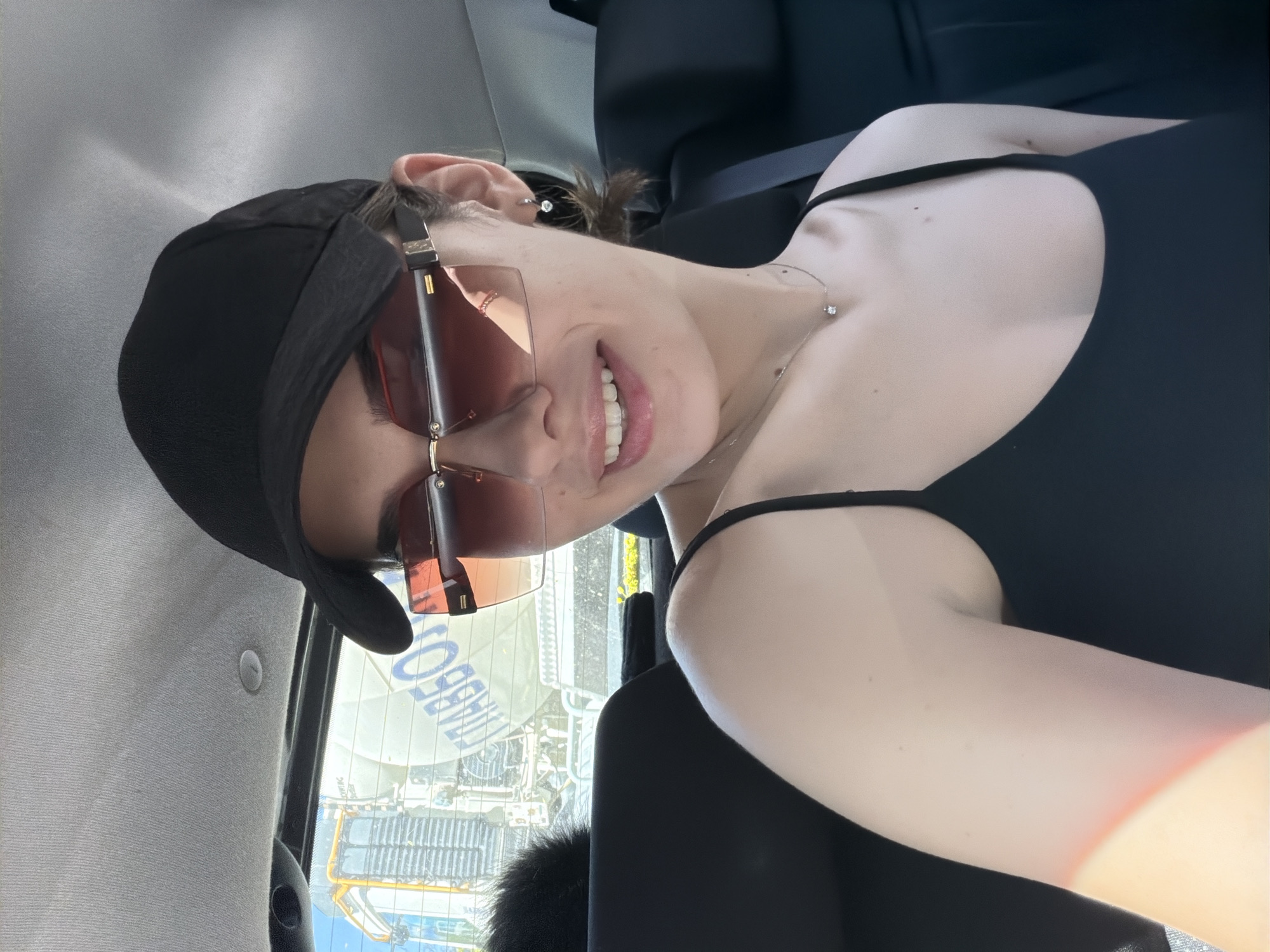 A young person wearing a black baseball cap, tinted sunglasses, and a black top is posing in a car. They are smiling and looking directly at the camera. A seatbelt is visible across their chest. Outside the car window, an urban scene in Sofia, Bulgaria, is visible. A large billboard with the word "AVEBECO" is prominent in the background, along with other buildings and city infrastructure.