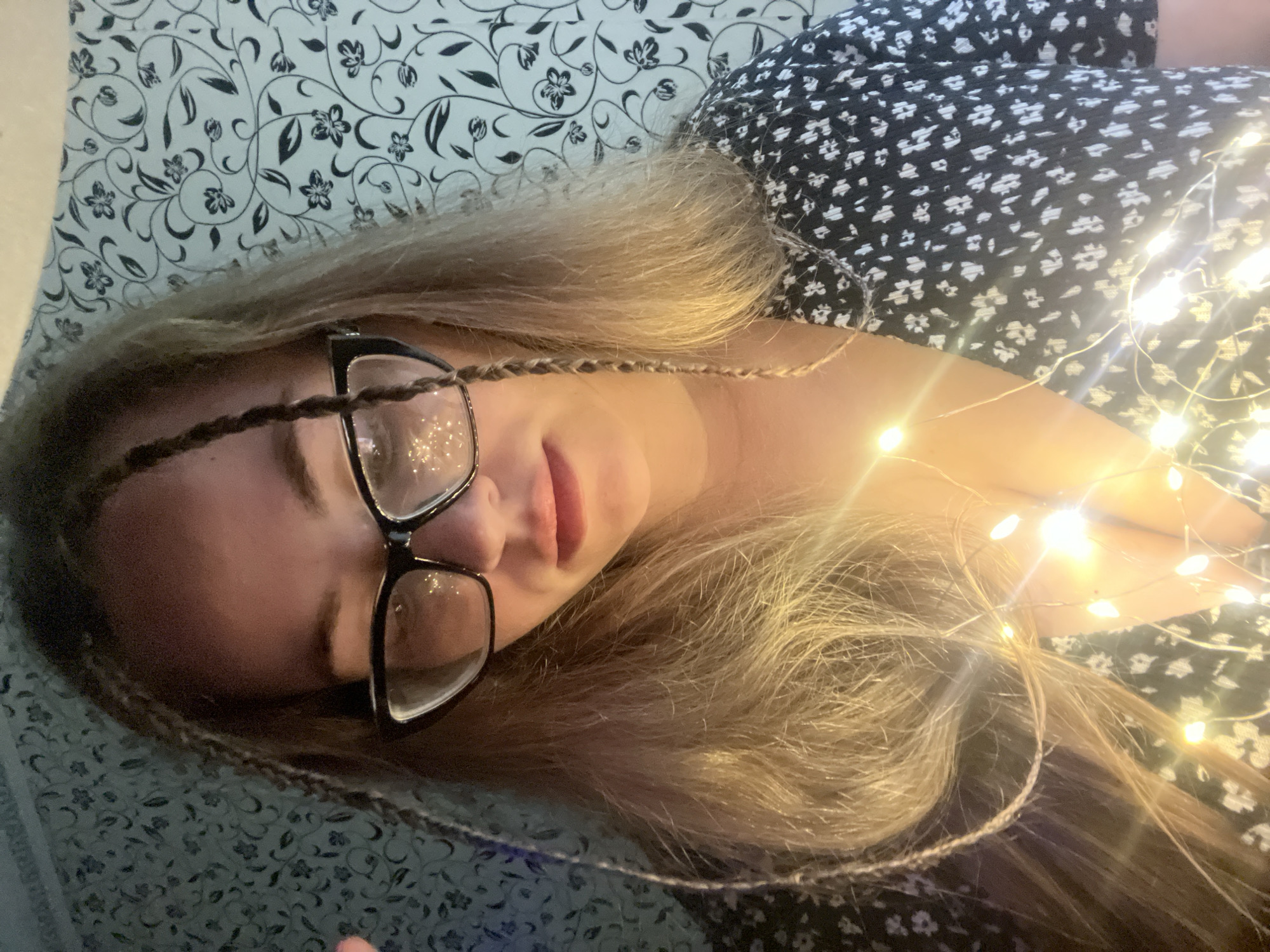 A woman lies on a floral patterned surface, possibly a bed, wearing a black and white dress. She wears glasses and has braids in her blonde hair. A string of fairy lights is draped across her body, illuminating her face and hair. There is no indication of the location being the city of Chaikovsky, Russia, in the image.