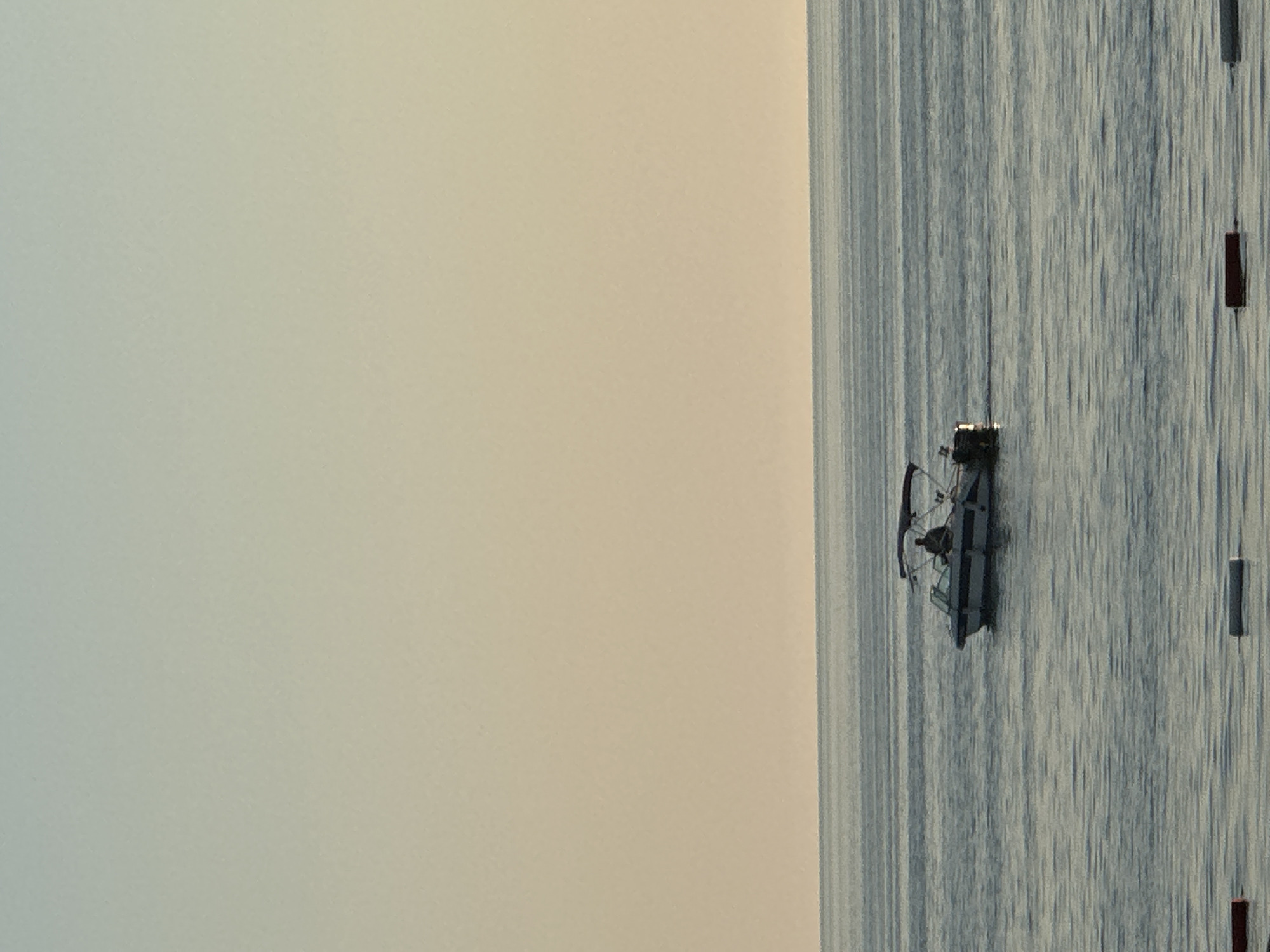 The image presents a serene coastal landscape, rotated 90 degrees counter-clockwise from its natural orientation. When viewed correctly, it depicts a tranquil body of water under an expansive sky, likely the Adriatic Sea near Bar, Montenegro.
A small, dark-hulled boat is positioned towards the right side of the frame, resting gently on the calm water. Its details are somewhat indistinct but suggest a fishing vessel or a small recreational boat, possibly featuring a canopy or cabin structure. Scattered further to the right on the water are several dark, elongated vertical objects, resembling buoys or markers, which could indicate a fishing net, a designated area, or a navigation route.
The water surface is remarkably smooth and reflective, displaying subtle ripples and soft, elongated textures caused by the ambient light. The sky, which fills the upper portion of the image, exhibits a delicate color gradient, transitioning from a pale, cool blue-green hue on the left to a warmer, creamy beige on the right. This diffused lighting suggests either the early morning glow of dawn or the soft light of dusk.
The overall atmosphere of the scene is one of peace and quiet, characterized by muted tones and clear, calm weather conditions. There are no individuals visible in the image, and no discernible text.