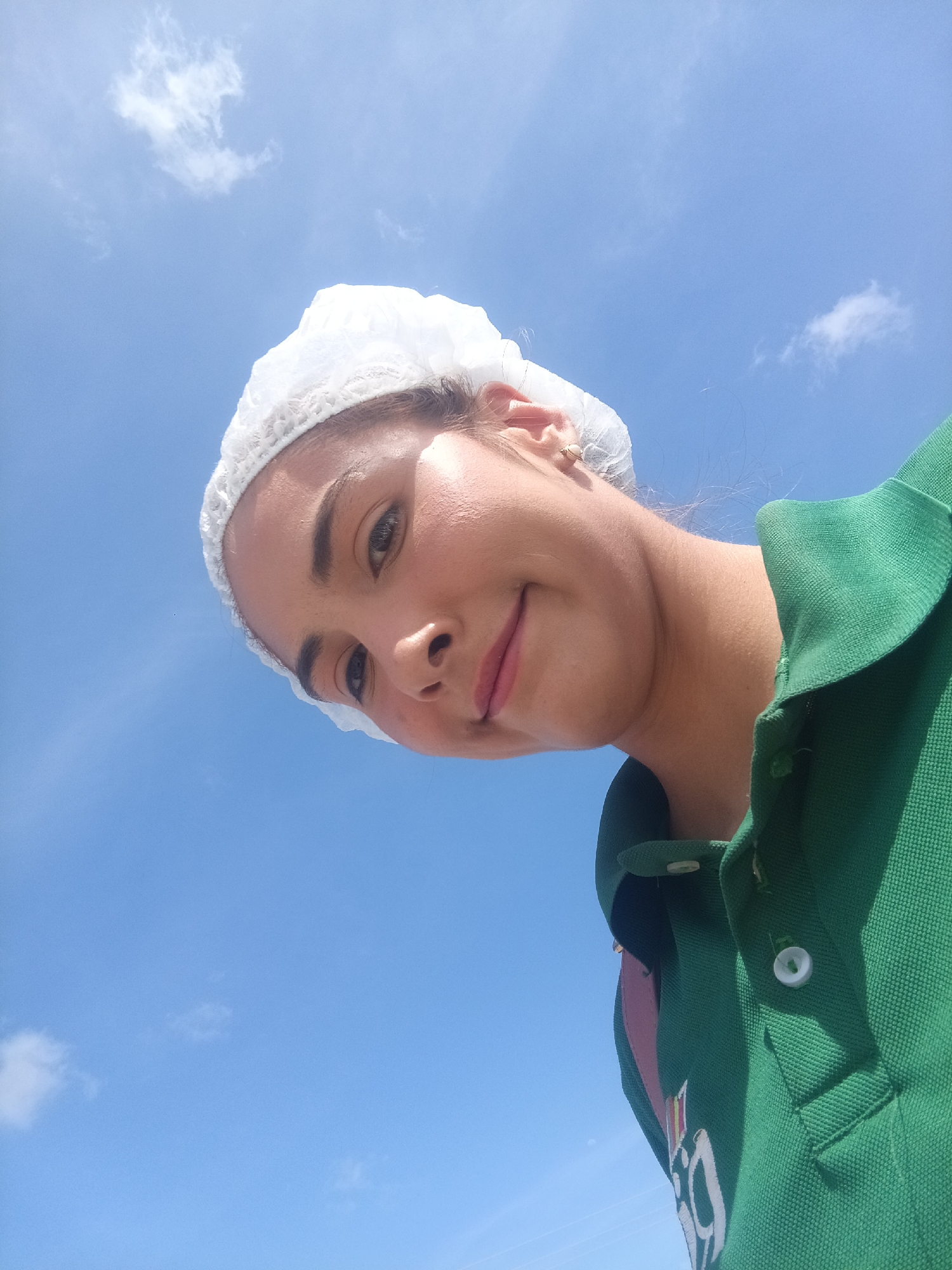 A woman wearing a white hairnet and a green polo shirt looks down at the camera from a low angle. The background is a bright blue sky with scattered white clouds. The woman appears to be smiling slightly. The image provides no information about Juan Griego, Venezuela.
