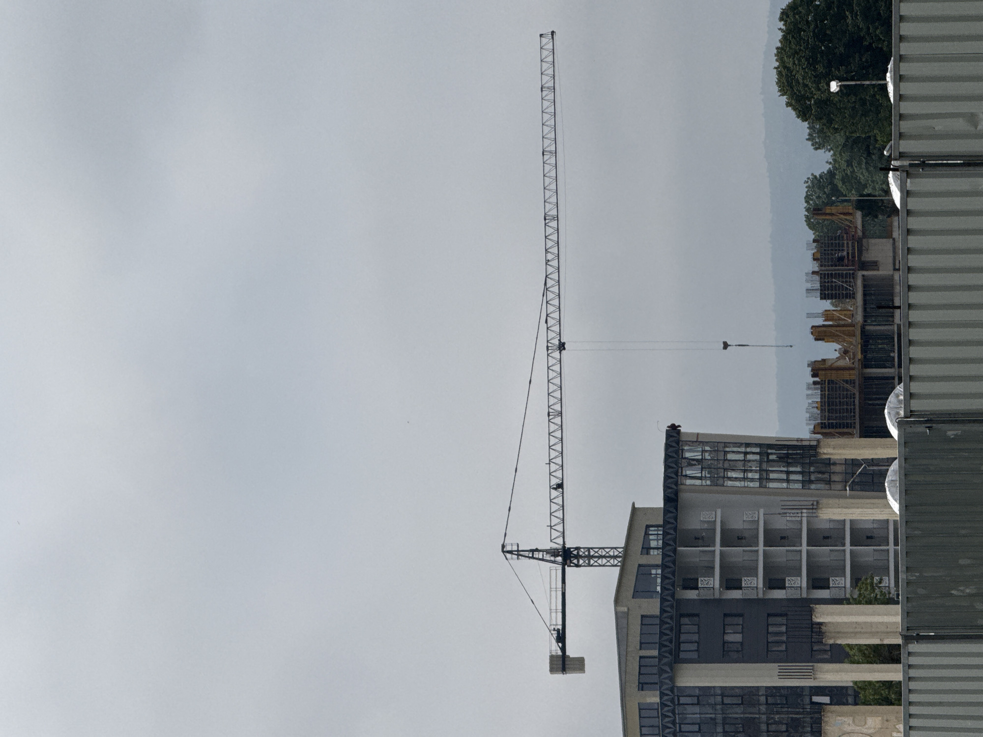 A landscape-oriented photograph depicts a construction site under an overcast sky in Varna, Bulgaria. The image is dominated by a tall, slender tower crane positioned on the left side, its lattice mast rising vertically and its horizontal jib extending to the right over a multi-story building under construction. The crane's hook and cable hang downwards, with a small object at the end.
In the mid-ground, a modern building is actively being constructed. Its concrete frame and partially installed dark-paned windows are visible, along with multiple uniform square or rectangular openings that appear to be balconies or window recesses on the upper floors. Some sections of the building are dark grey, while others are lighter, possibly concrete or a light-colored facade material.
To the far right, a stack of grey, corrugated shipping containers or modular site offices forms a solid barrier, partially obscuring the view of the construction area behind it. Beyond the immediate construction site, in the background, a dense line of green trees suggests a wooded area or hillside, fading into a hazy, lighter-toned distant landscape or horizon.
The sky is a uniform, light grey, indicating an overcast or cloudy day with diffused lighting. No direct sunlight is visible, and the overall mood is somewhat muted. There are no clearly discernible people or visible text in the frame. The scene captures an active phase of urban development.