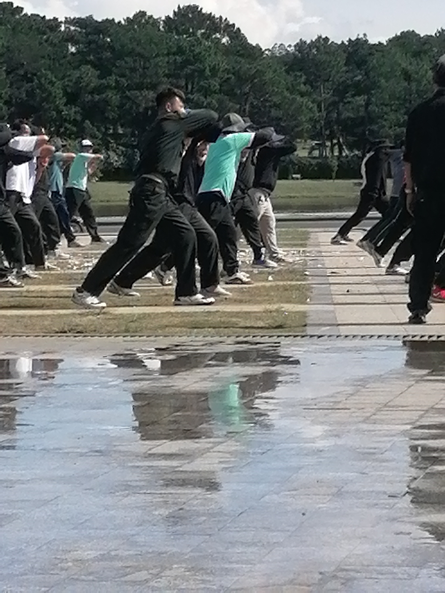 The image captures a group of individuals performing what appears to be a martial arts or synchronized exercise routine outdoors, possibly in Da Lat, Vietnam, given the lush green trees in the background and the paved, reflective ground. The individuals are dressed in athletic attire, with several wearing dark long-sleeved shirts and dark pants, while others are in lighter colored shirts, including a prominent teal shirt. Their stances and arm movements suggest a coordinated activity. The ground is wet and reflective, indicating recent rain or a water feature, with puddles and reflections of the sky and trees visible. The environment appears to be a public space with a tiled or paved walkway and a grassy area beyond, lined with dense trees. The lighting suggests it is daytime with overcast skies.