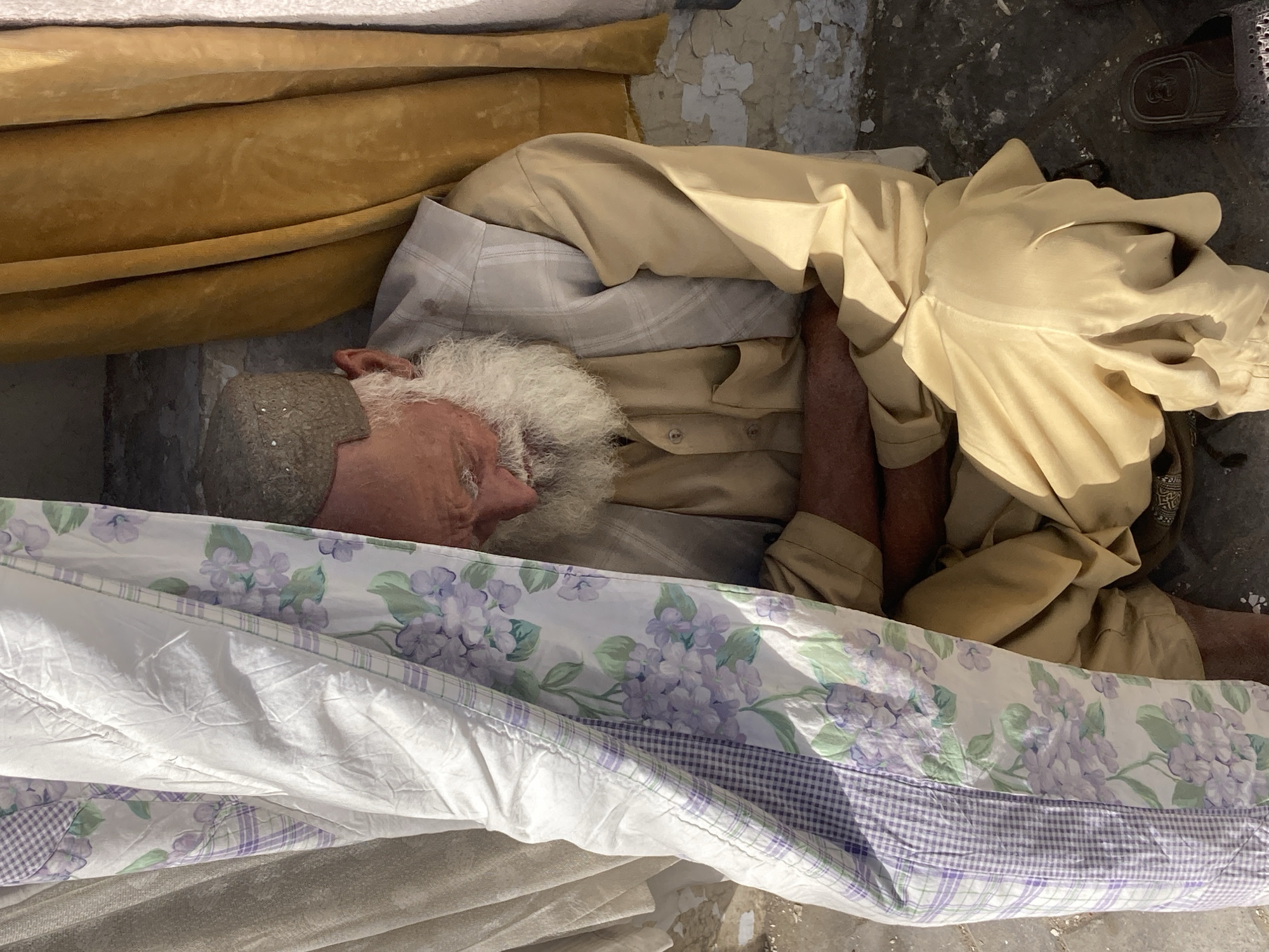 A deceased elderly man with white hair and beard lies in an open coffin in Quetta City Tehsil, Pakistan. He is dressed in a tan shirt and plaid pajama pants. The coffin is draped with a white blanket decorated with purple flowers. A tan fabric lines the inside of the coffin. The floor is concrete with flaking paint. A brown shoe is visible in the upper right corner.
