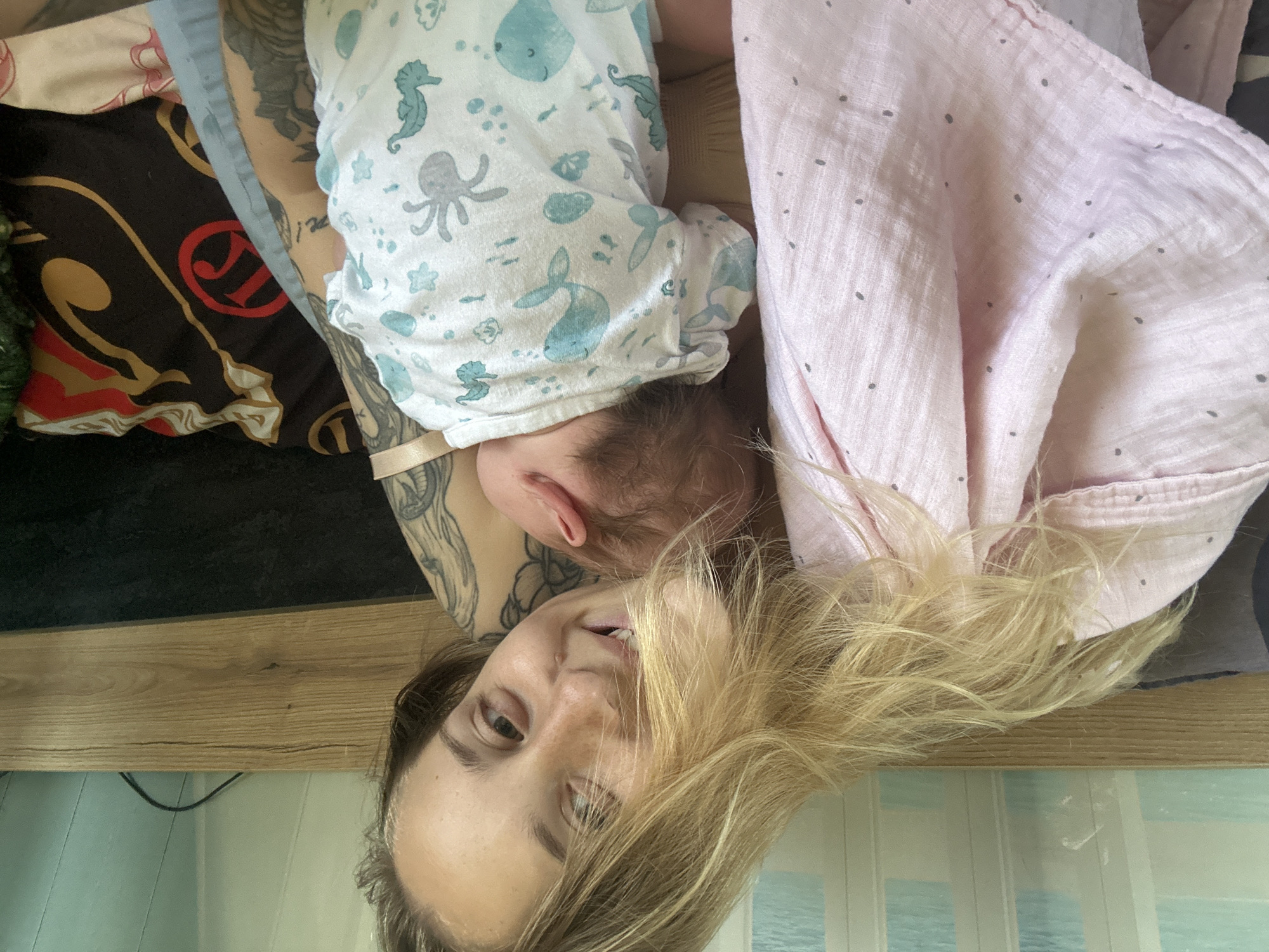 A young Caucasian woman with blonde hair and tattoos on her arm is holding a newborn baby. The baby is wearing a white onesie with blue sea creature prints. The woman is smiling and looking at the camera. They are lying down on a bed with a wooden headboard. A pink blanket with grey dots is draped over the woman's shoulder. The background shows a wall with a patterned wallpaper and a black pillow with red and gold designs. The scene is indoors, likely a bedroom.