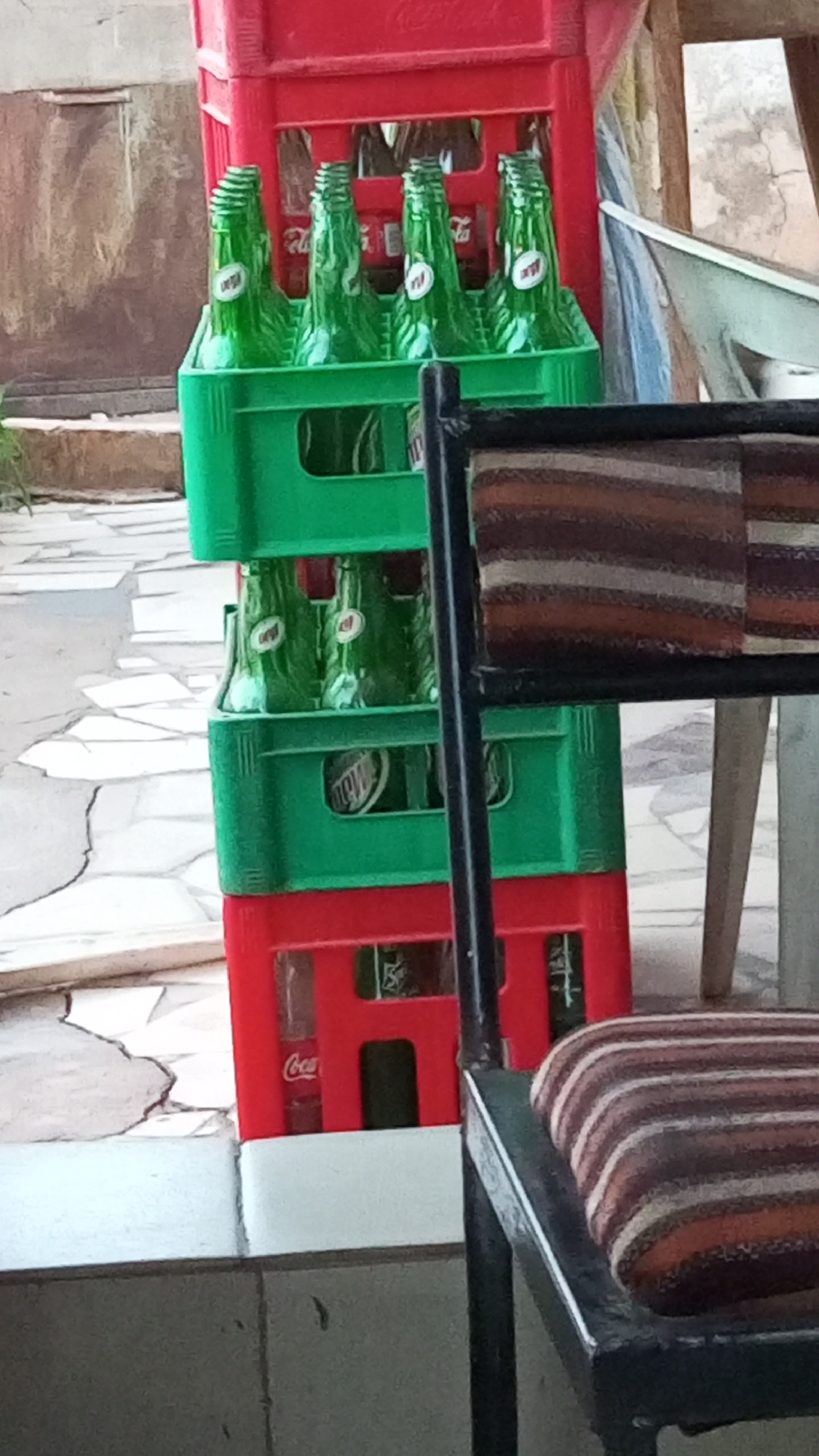 A stack of plastic crates containing glass bottles, likely soft drinks, is visible outdoors in Sabon Tasha, Nigeria. The crates are stacked in alternating colors of red and green. The green crates hold green glass bottles labeled "Mountain Dew," while the red crates contain Coca-Cola bottles. A chair with a striped cushion is partially in the foreground. The background shows parts of a tiled floor and walls of a building.