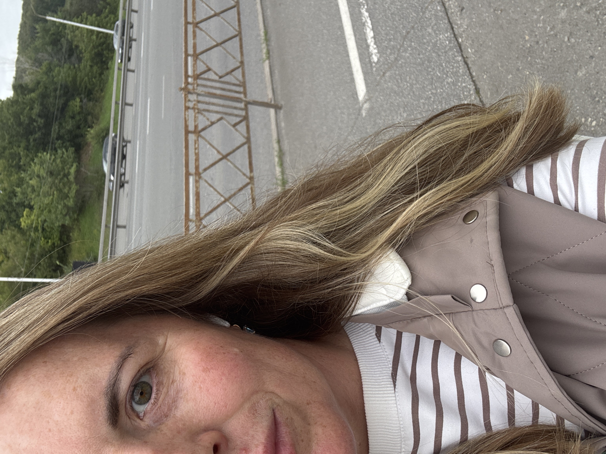 A partial, close-up, outdoor, eye-level, medium shot shows the left side of a fair-skinned woman's face, her blonde hair, and her torso, with a blurred background of a highway and trees. The woman is wearing a light-colored collared jacket over a horizontally striped long-sleeved shirt. Her hair is light brown with blonde highlights, and it frames her face. She has green eyes, and is looking slightly to the left. In the background, two dark-colored cars are visible on a highway, and trees and greenery line the road. The scene appears to be in Ufa, Russia, as indicated by the presence of a typical Russian guardrail separating the sidewalk from the highway.