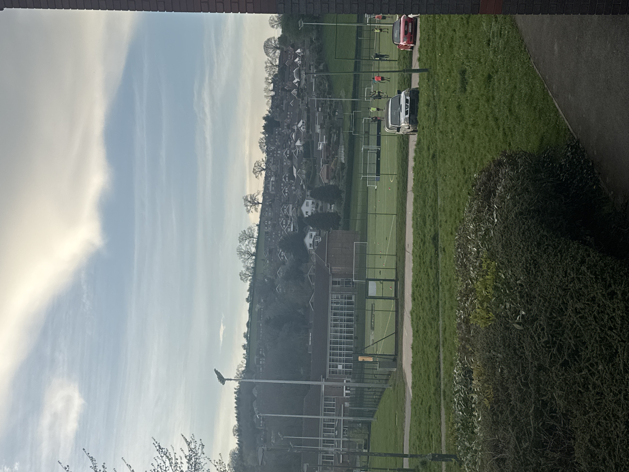 The image shows a view from a window, likely in Welshpool, United Kingdom, as indicated by the presence of football/sports pitches in the background. The sky is predominantly blue with wispy clouds. In the middle ground, there is a cluster of buildings, possibly houses, on a slight hill. Further in the background, sports pitches are visible, with people appearing to play or engage in activities. Vehicles, including a red van or car and a white van, are parked alongside the pitches. The immediate foreground is occupied by a hedge or bushes. The top and bottom of the image are framed by a brick wall or other building structure, suggesting an indoor viewpoint.
