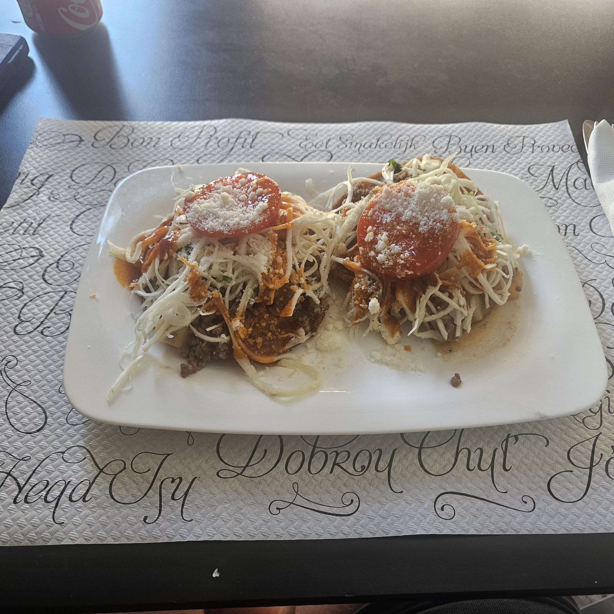 The image shows a white rectangular plate with two circular food items topped with shredded cheese, sliced tomatoes, and a red sauce, resting on a patterned placemat. The placemat features black script text that reads "Bon Profit" and "Dobroy Chut". The food appears to be some type of savory pastry or flatbread. In the background, a portion of a red and white Coca-Cola can and a dark surface, possibly a table, are visible. A metallic utensil is also partially visible to the right of the plate. The overall setting suggests a dining environment. Without further information, the specific location within Girona, Spain cannot be determined from the visual cues alone.
