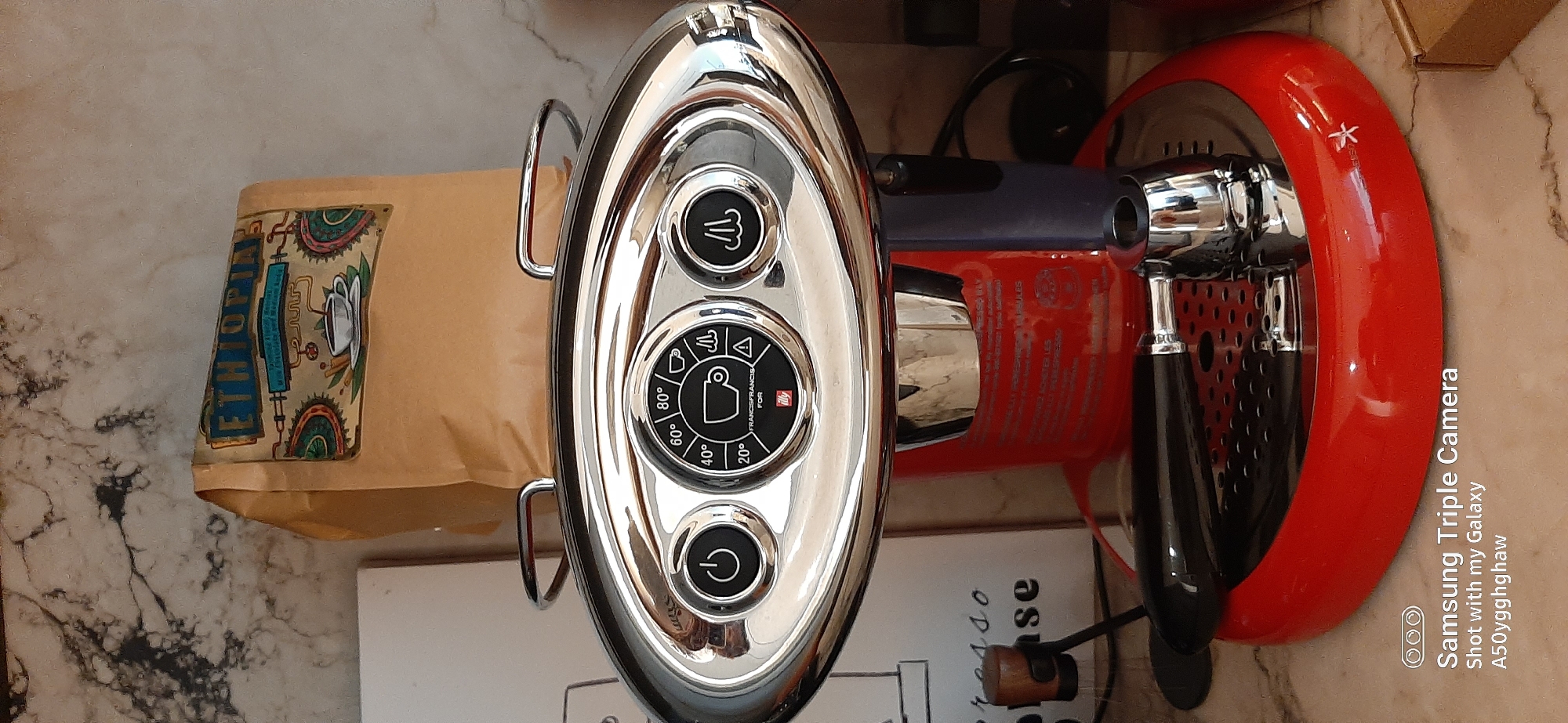 This media file displays a close-up, top-down shot of a coffee preparation setup on a marble-patterned surface. The main subjects are a red and chrome espresso machine and a brown paper bag of coffee.
The coffee machine, a sleek Illy FrancisFrancis! model, features a shiny chrome oval front panel with three circular buttons. The top button has a steam icon, the bottom a power icon. The central button acts as a dial, displaying temperature settings (20°, 40°, 60°, 80°), coffee cup icons, a steam icon, a warning triangle, and the text "FRANCISFRANCIS FOR Illy". The machine's body is a vibrant red, with a dark portafilter handle visible on the right.
Adjacent to the machine, a brown paper bag of coffee stands upright. Its colorful label prominently reads "ETHIOPIA," indicating the coffee's origin, and features an intricate illustration, possibly depicting coffee plants or processing equipment.
Partial text visible on a white object below the machine reads "esso," likely part of "espresso." A watermark in the bottom right corner identifies the image as "Samsung Triple Camera Shot with my Galaxy A50ygghghaw." The scene is illuminated by indoor lighting, typical of a kitchen or cafe setting. The location context for this setup is Dubai, United Arab Emirates.