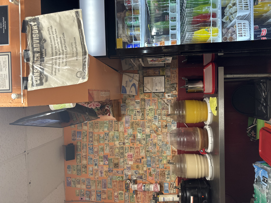 This image depicts the interior of a commercial food establishment located in Coral Springs, United States of America. The primary visual is an orange wall extensively covered with various international banknotes, forming a decorative display. A black flat-screen television is mounted at an angle on this wall, above a small shelf with pink artificial flowers. A black speaker is also affixed to the orange wall.
An overhead orange beam features a white "CONSUMER ADVISORY" sign, secured with black tape, which outlines risks associated with consuming raw or undercooked meats, poultry, seafood, or eggs, and references "FreshFromFlorida.com." Below, framed certificates, including one labeled "ServeSafe Certification," are visible on the wall. To the far left, a white utility dispenser is mounted on the wall.
In the foreground, a stainless steel counter holds three large, clear plastic beverage dispensers on white circular bases, containing opaque cream, translucent reddish-brown, and bright yellow liquids, respectively. To the left of these dispensers, a black electric warmer or pot is visible. To their right, a metal caddy containing utensils, a stack of red and black serving trays, and a yellow cloth are arranged on the counter.
On the far right, a tall, glass-fronted commercial refrigerator is partially visible, stocked with multiple shelves of bottled and canned beverages, including colorful Jarritos sodas, Coca-Cola products, and other soft drinks. A lower shelf in the refrigerator holds white baskets containing numerous small, lidded plastic containers. The ceiling consists of white acoustic tiles. No individuals are present in the scene.