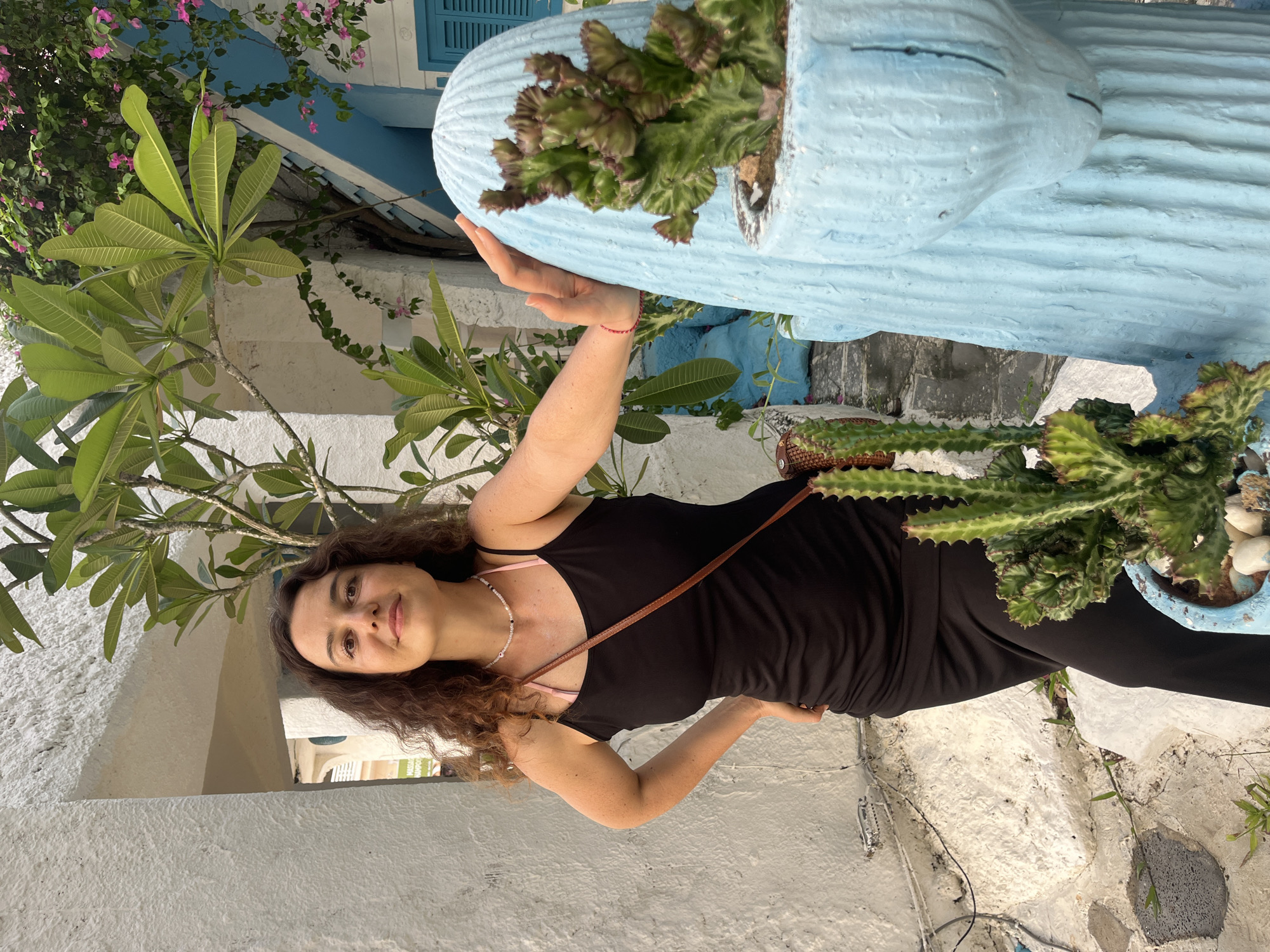 A woman with curly brown hair and dark clothing poses in front of a white wall. She is wearing a black tank top with a pink bra strap visible, a beaded necklace, and a brown strap across her chest. She is holding a light blue, textured pot that contains a succulent plant with green and reddish-brown leaves. The pot is shaped like a face with closed eyes and a closed mouth. To her left, a leafy green plant with large leaves grows. Behind her, there is a white wall, a blue-shuttered window, and a flowering pink plant. The scene appears to be outdoors in daylight, possibly in a tropical setting given the plants and the warm lighting. The woman is smiling faintly and looking towards the camera, appearing relaxed and content. The ground beneath is rough and textured with white and gray stones and some greenery. The overall impression is a casual outdoor portrait in a visually interesting location.
