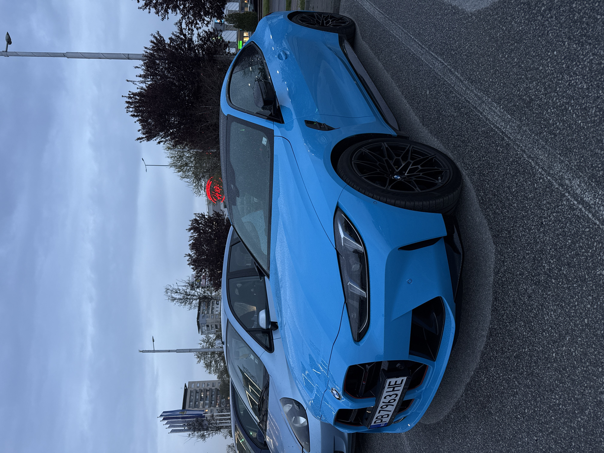 A blue BMW M3 G80 is parked on an asphalt surface in an urban area. The car has a black grille and black rims. The license plate reads "PB7963HE," which is a Bulgarian license plate. The photograph appears to be taken on an overcast day, possibly after rain, as there are water droplets on the car. In the background, there are trees, buildings, street lights, and a red neon sign. The location is Plovdiv, Bulgaria, based on the Bulgarian license plate.