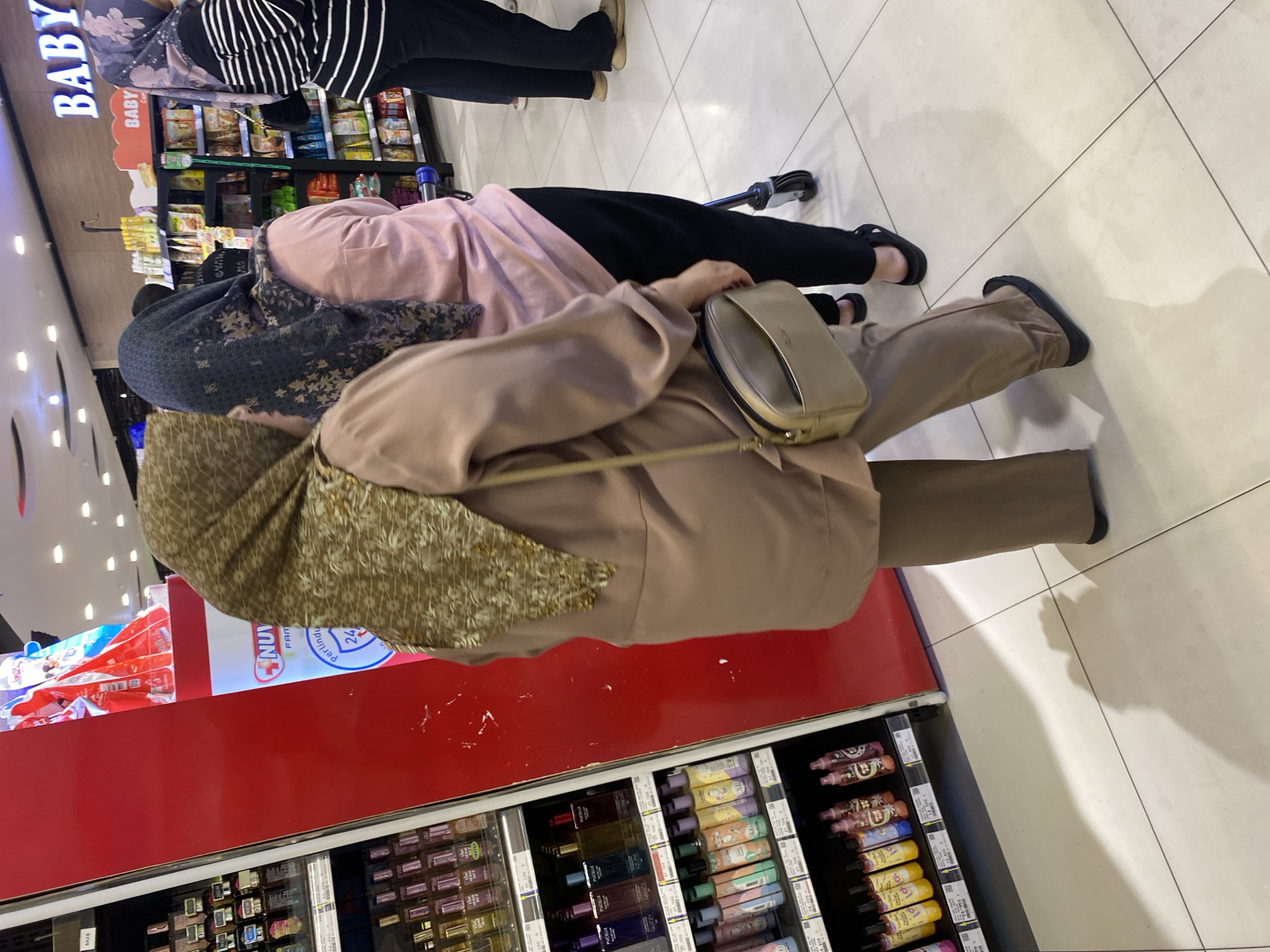 In an indoor retail environment, likely a department store or supermarket aisle in Banda Aceh, Indonesia, three individuals are visible. The foreground is occupied by a person wearing a tan hijab and a matching tan tunic and trousers, carrying a small, light-colored shoulder bag. To their left, a retail display features rows of colorful bottles, possibly perfumes or body sprays, with prices listed. To the right of the main subject, another person is partially visible, wearing a dark hijab with a floral pattern and a pink t-shirt. Further to the right, a third person, seen from the waist down, is dressed in a black and white striped top and black trousers, wearing black sandals. They are standing near a shopping cart with a blue handle. The environment includes illuminated ceiling lights and a sign with the word "BABY" prominently displayed. The floor is tiled with light-colored squares.