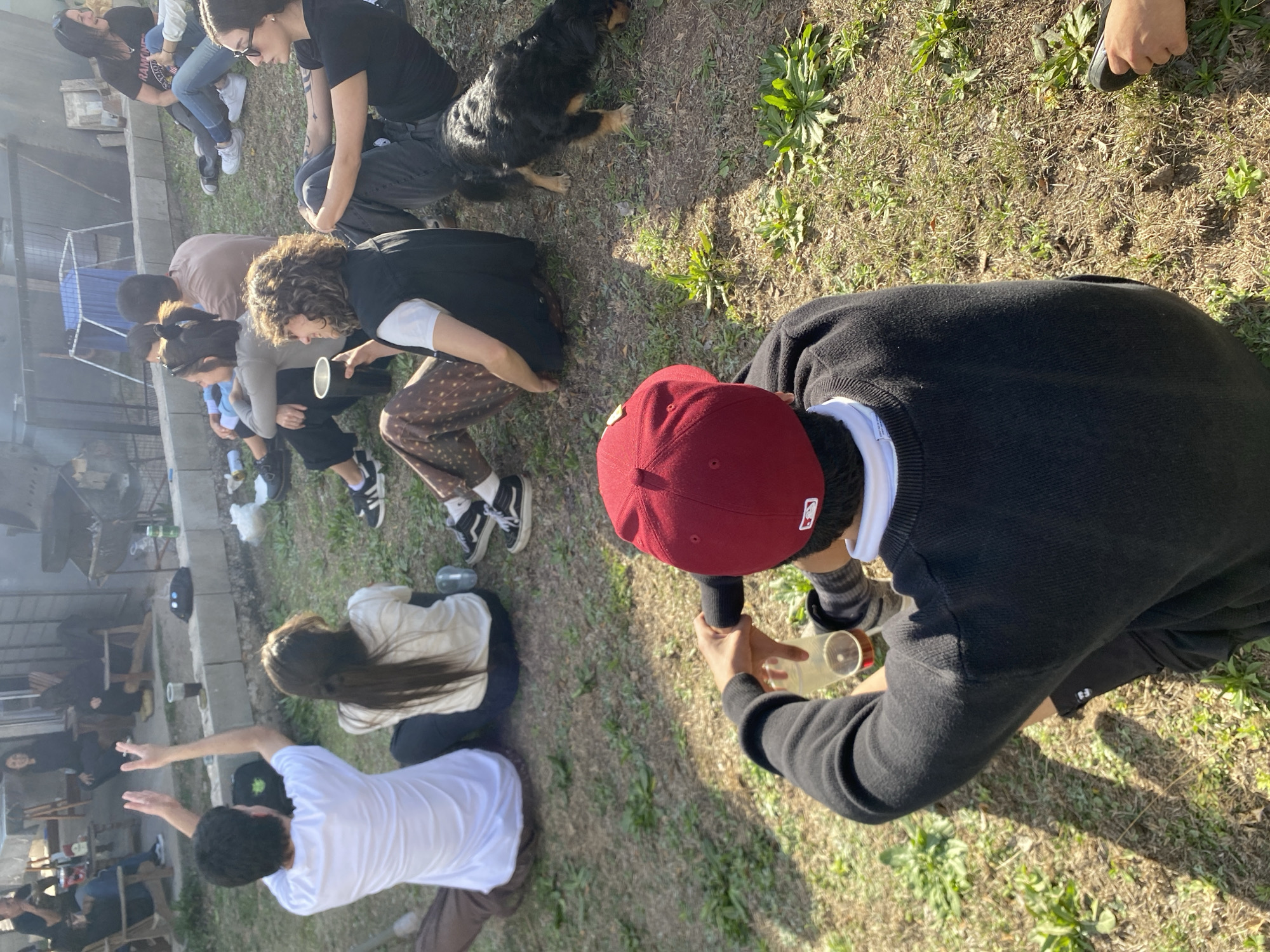 In the city of Comuna Estancia Vieja, Argentina, on a grassy outdoor area with scattered patches of dry earth and sparse green vegetation, a group of people are gathered. In the foreground, a person wearing a red baseball cap and a black sweater sits on the ground, holding a clear plastic cup with a beverage. Behind them, other individuals are seated on the grass. To the right of the foreground subject, a medium-sized dog with black and brown fur is sniffing the ground. In the background, a table with some items on it and a grill are visible, suggesting a casual outdoor gathering. The overall environment appears to be a backyard or a park area.