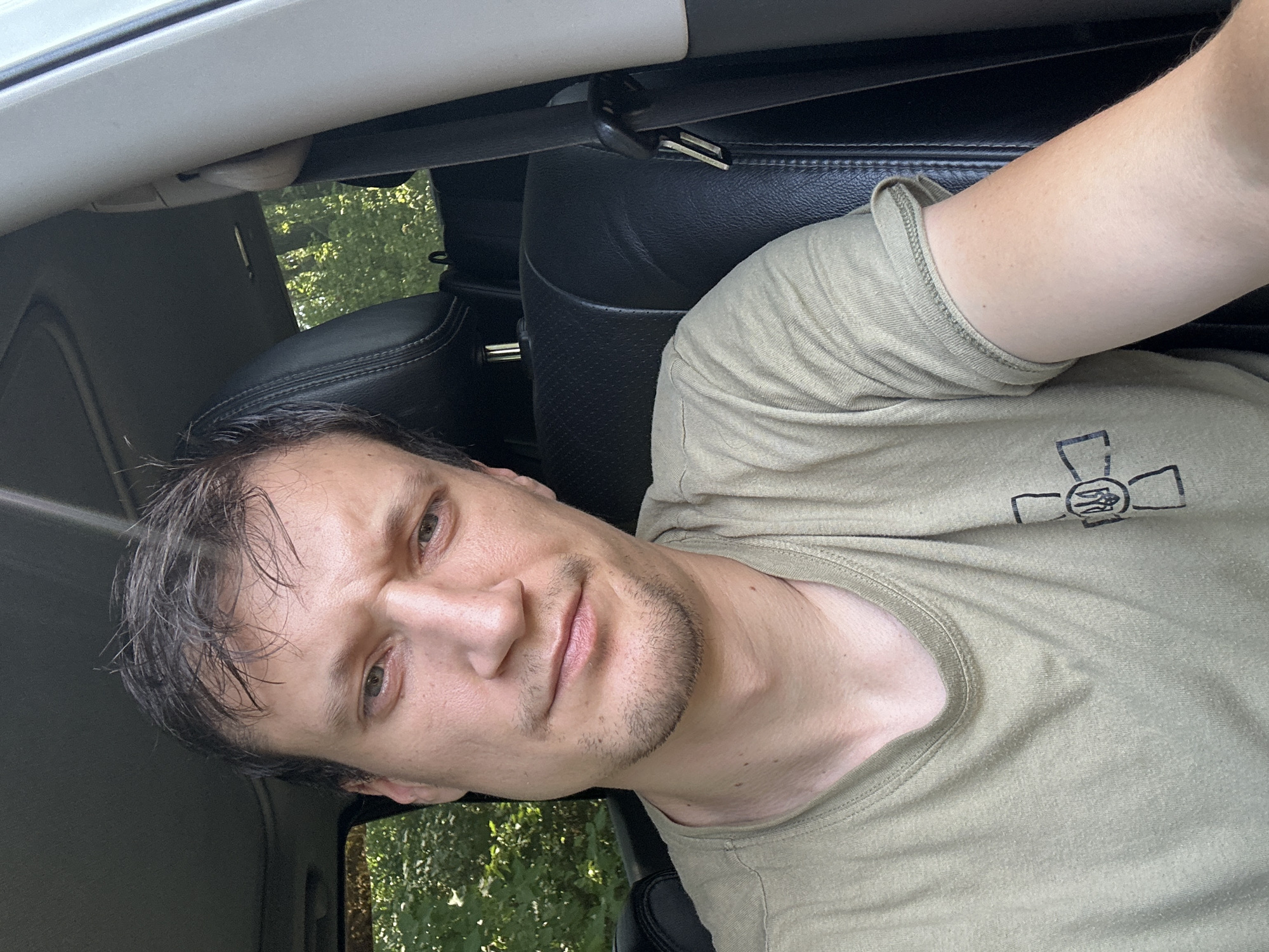 A man is sitting in the driver's seat of a car. He is wearing a light green t-shirt with a black emblem on the left side of his chest. The emblem appears to be a cross with a trident inside it. The man has short, dark brown hair and light-colored eyes. He is looking towards the camera with a slight frown. The interior of the car is visible, including black leather seats and a seatbelt. Through the windows, trees and greenery can be seen, suggesting an outdoor, possibly rural, location. The scene is illuminated by natural light.