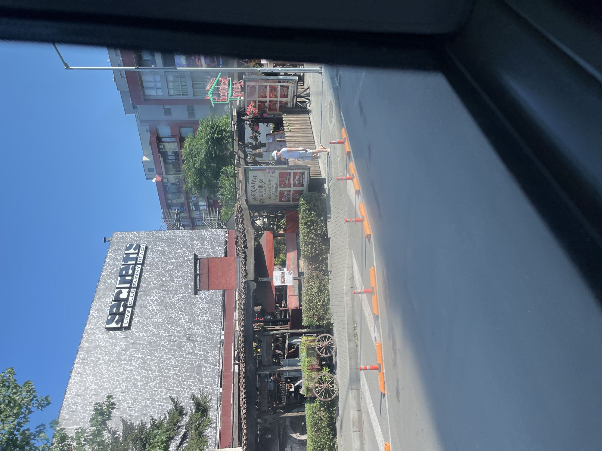 This is a street-level photograph taken in Nesebar, Bulgaria, likely during daylight hours given the clear blue sky and bright sunlight casting shadows. The viewpoint appears to be from a moving vehicle, indicated by the dark frame in the lower right corner.
The dominant structure in the background is a large, modern building with a facade covered in a patterned grey material. A prominent sign on this building reads "secrets" in large, illuminated letters, with smaller text below stating "LIVE CLUB SUNNY BEACH". Adjacent to this modern building is a more traditional, rustic structure with a wooden roof supported by posts, creating a covered outdoor seating area or facade for a restaurant. This structure has an old-fashioned wooden cart with large wheels displayed in front of it, filled with greenery. Signs displaying food menus with images are affixed to the wooden fence and walls of this establishment.
A person, appearing to be a woman wearing a white cap, white t-shirt, and light-colored shorts, is walking on the sidewalk in front of the traditional establishment. She is facing away from the camera. The street in the foreground is paved and has a white crosswalk marked. Orange traffic cones and barriers line the edge of the sidewalk, delineating the pedestrian area from the road. In the mid-ground, various trees and green bushes are visible, adding to the outdoor ambiance. Beyond the "secrets" building, other multi-story buildings with balconies and windows are visible, suggesting an urban or resort area. The overall impression is a street scene in a busy tourist destination, likely a resort town like Sunny Beach, which is near Nesebar.