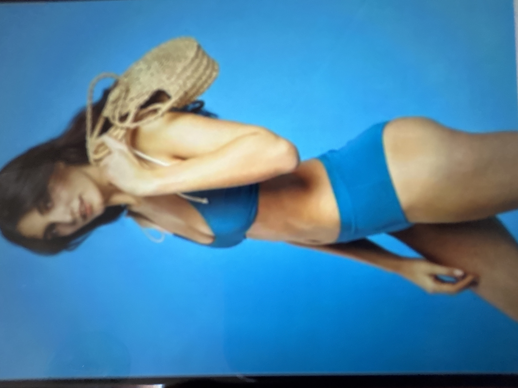 A woman wearing a blue bikini and a straw bag over her shoulder is posing against a blue background. Her body is contorted, with her head tilted back and her arms wrapped around herself. The lighting suggests a studio setting, with a bright, even illumination. The overall mood is one of leisure and relaxation, perhaps hinting at a vacation or beach setting. There is no visible text in the image. The context of Gelendzhik, Russia, is not discernible from the image itself.