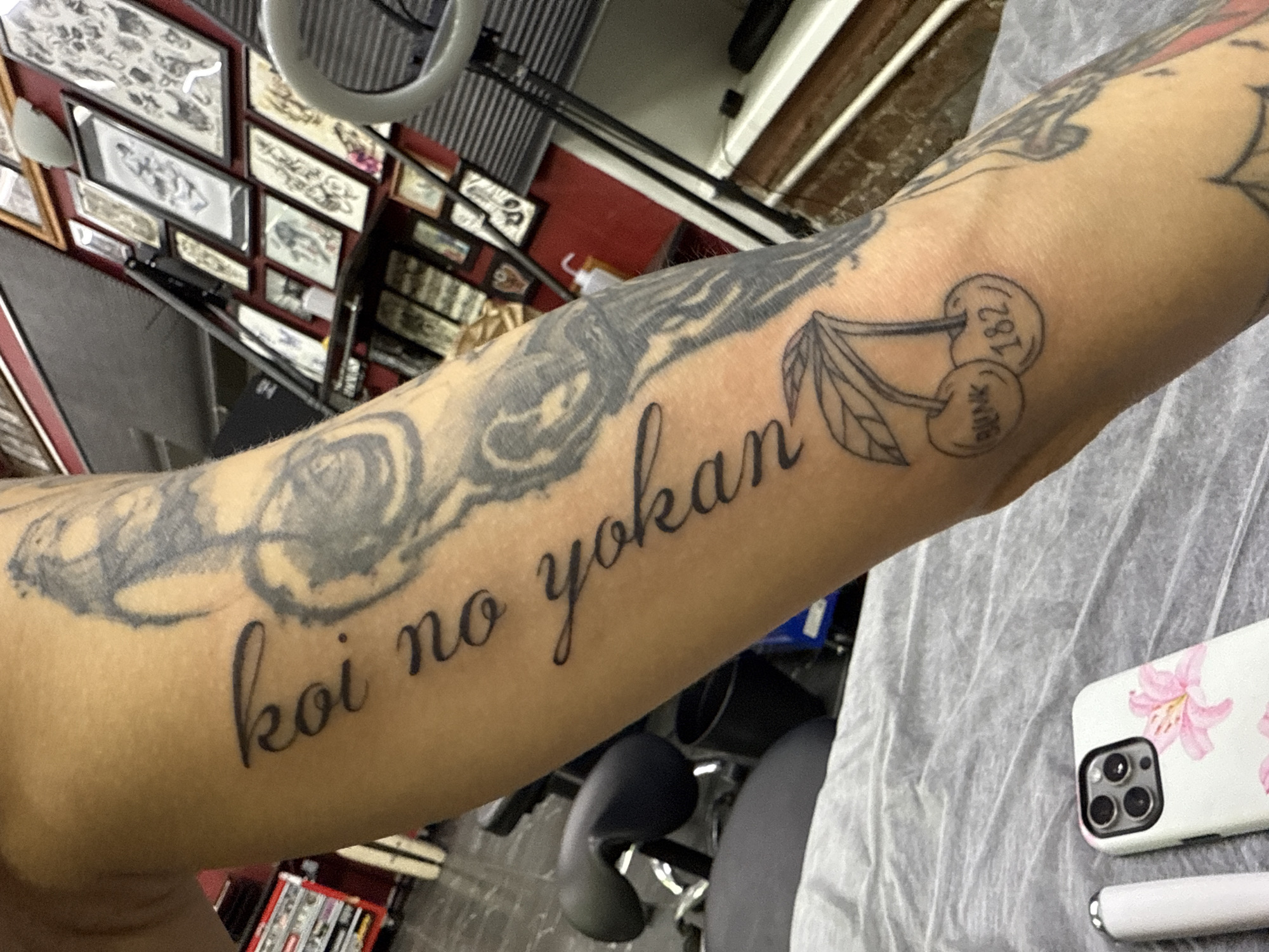 The image shows a person's arm with multiple tattoos. The tattoos include a grey tattoo of a rose with accompanying leaves. Another tattoo is a line drawing of two cherries; one cherry has the number "182" printed on it, and the other cherry has the word "BLINK" printed on it. Another tattoo is a phrase in black cursive, reading "koi no yokan". The person is sitting in a tattoo studio. A mobile phone with a floral case is also in the shot.